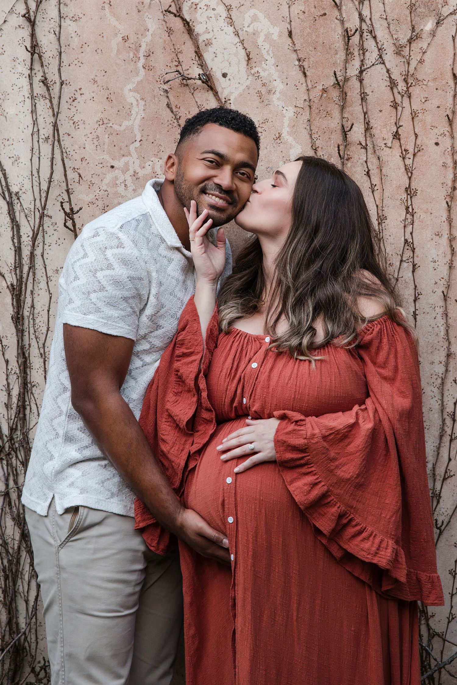 A pregnant woman in a red dress kisses her partner's cheek while he smiles and holds her baby bump.