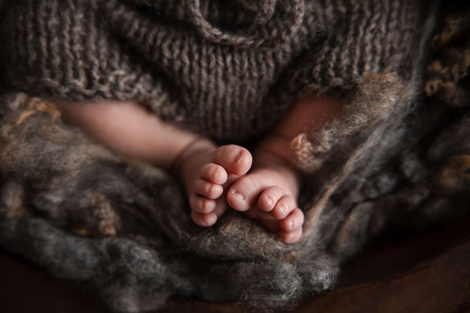 Tiny bare feet peek out from underneath a warm knitted gray blanket.