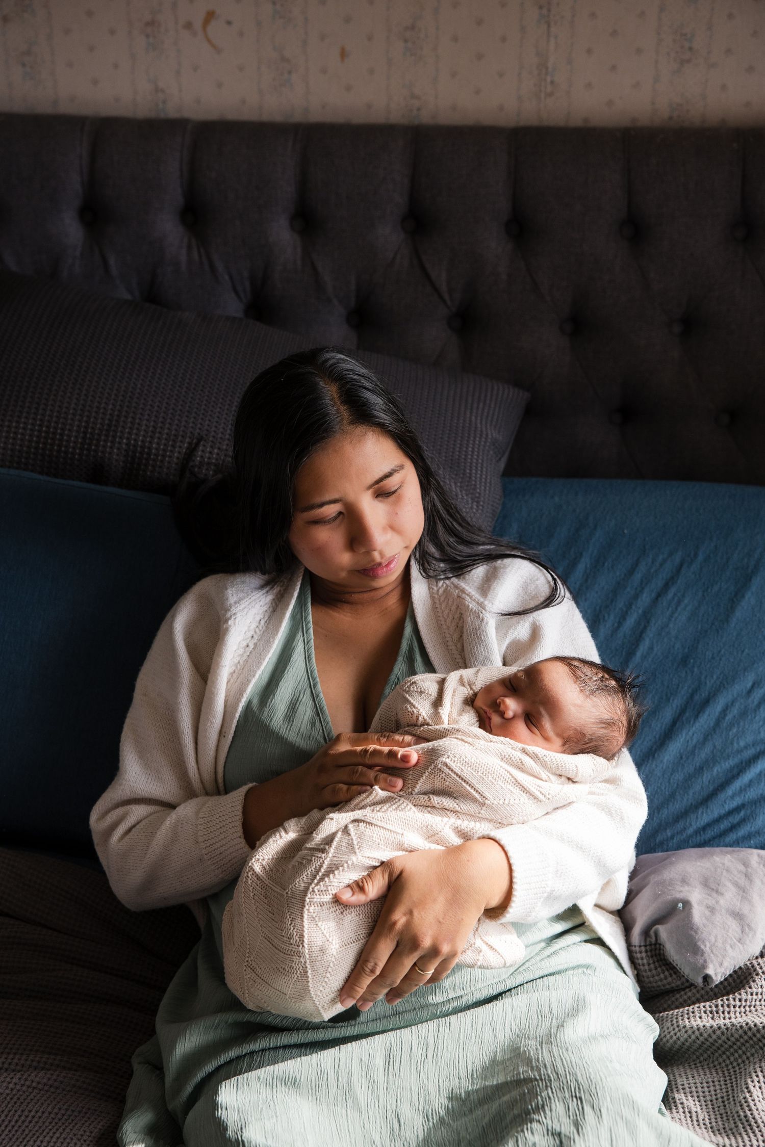 A mother in a white robe holds her sleeping newborn baby while resting on a bed with blue bedding.