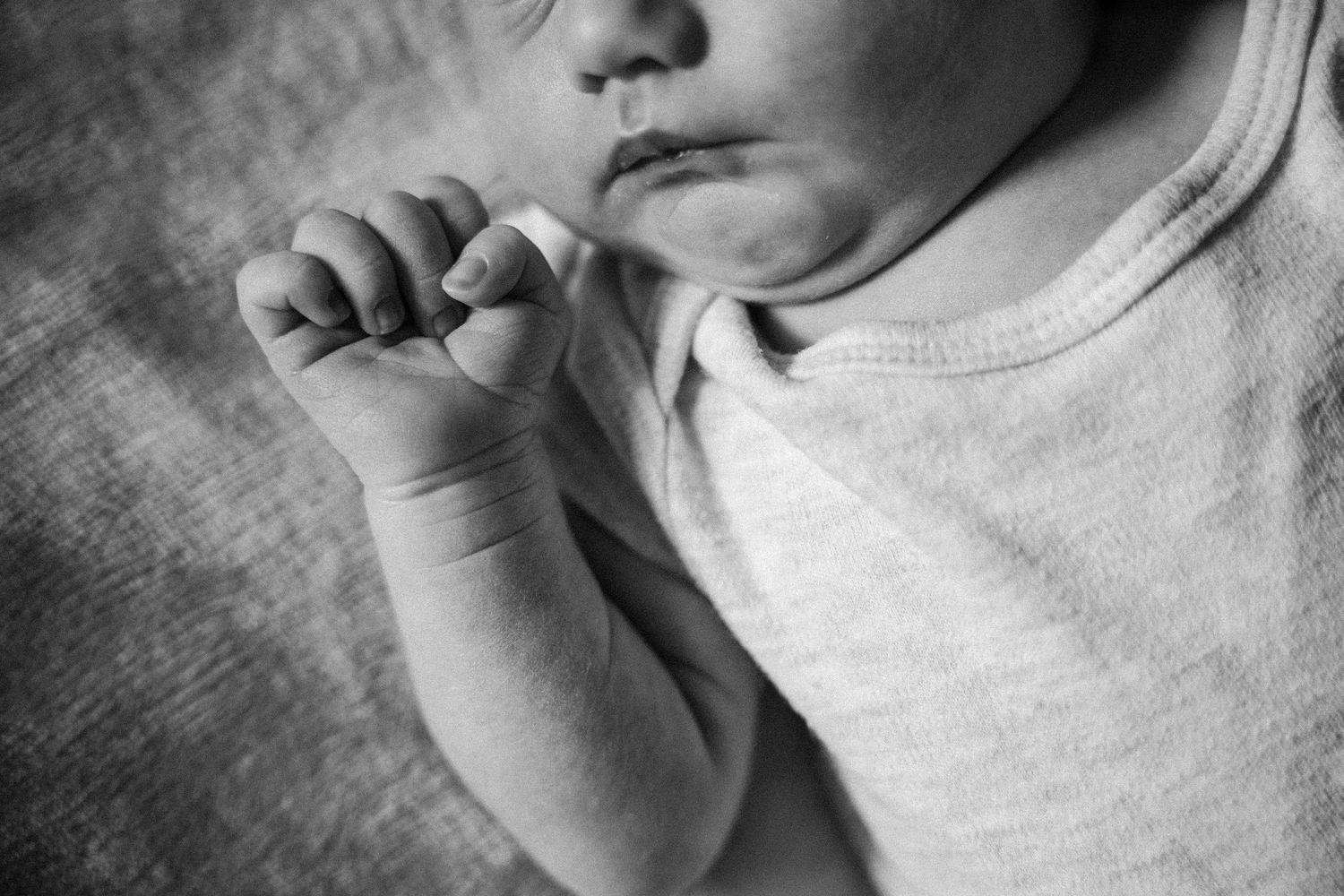 Black and white photo of a sleeping newborn baby in a white onesie, gently holding their tiny fist near their mouth.