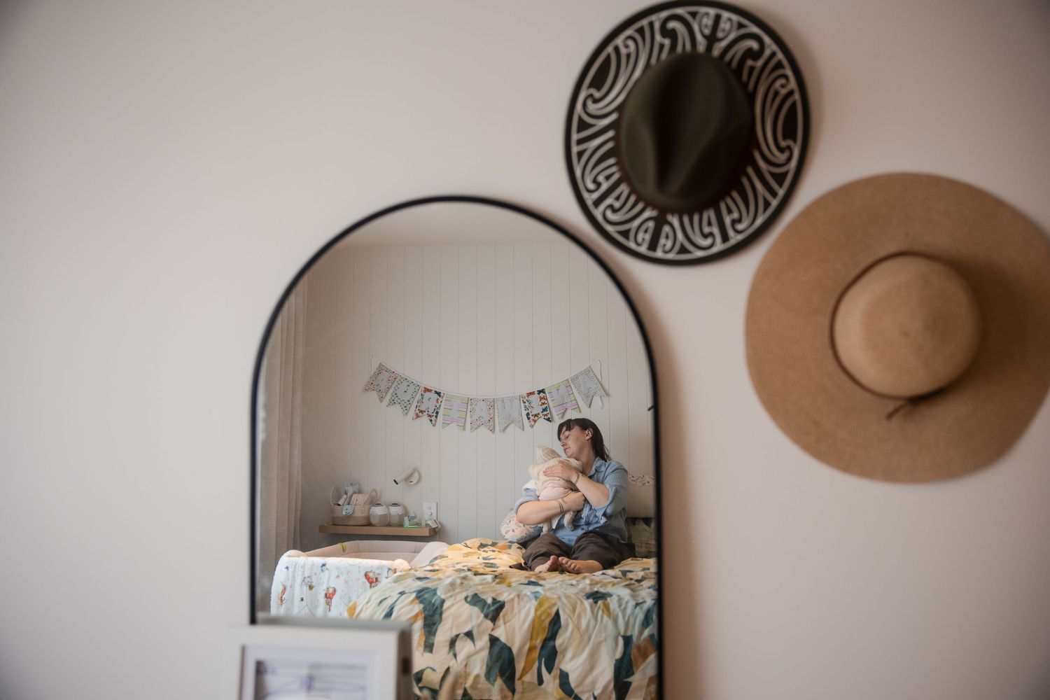 Woman holding newborn baby reflected in arched mirror with straw hat wall decor in cozy nursery bedroom.