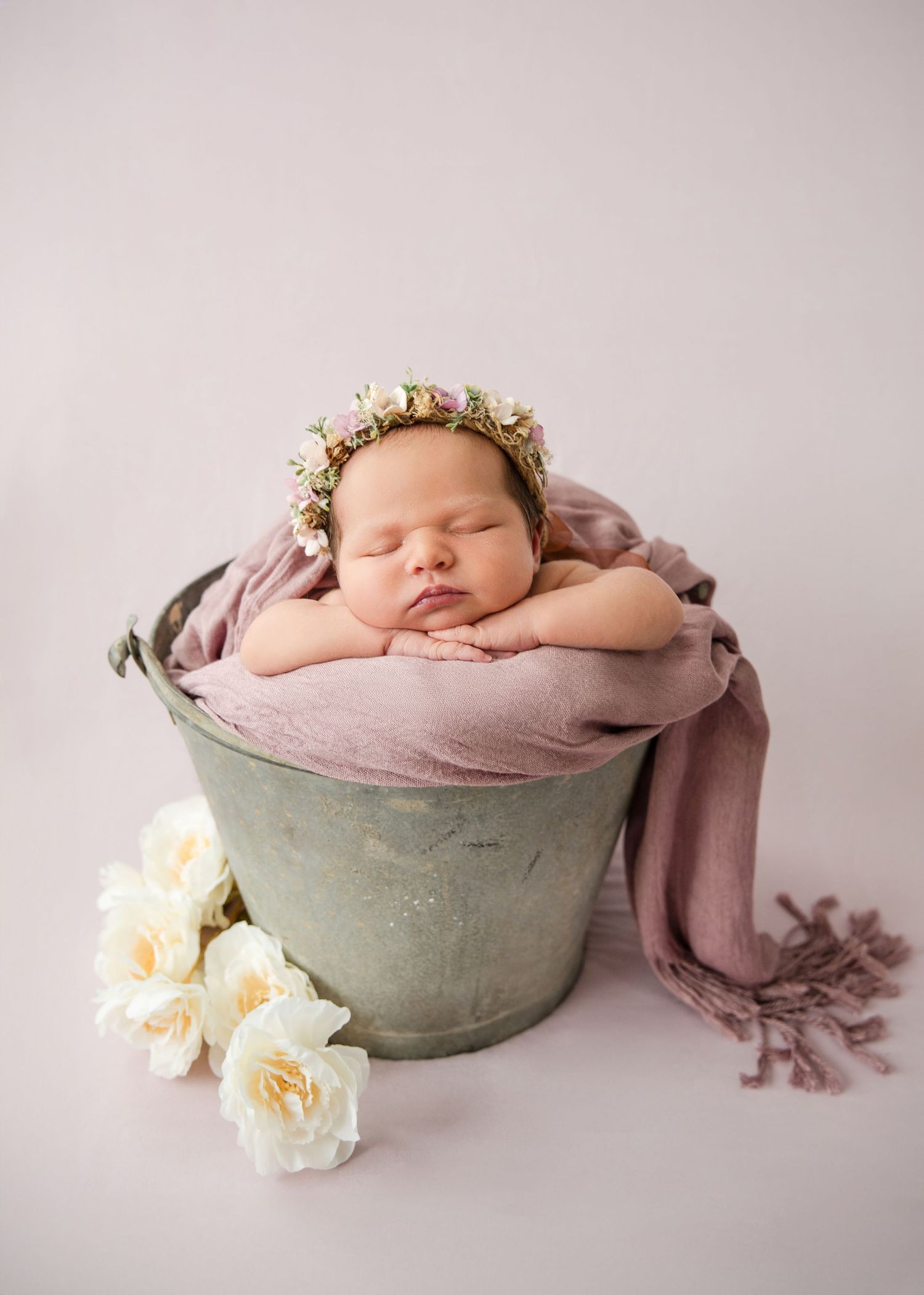 Sleeping newborn wrapped in pink fabric rests in vintage metal bucket with white roses beside it.