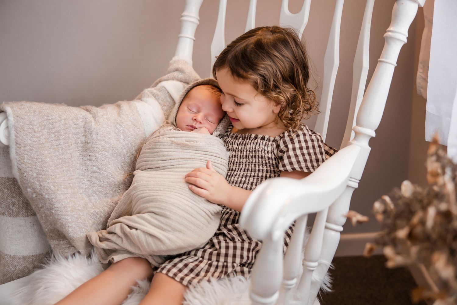 Toddler girl with curly hair gently holding swaddled newborn baby sibling in a white rocking chair.