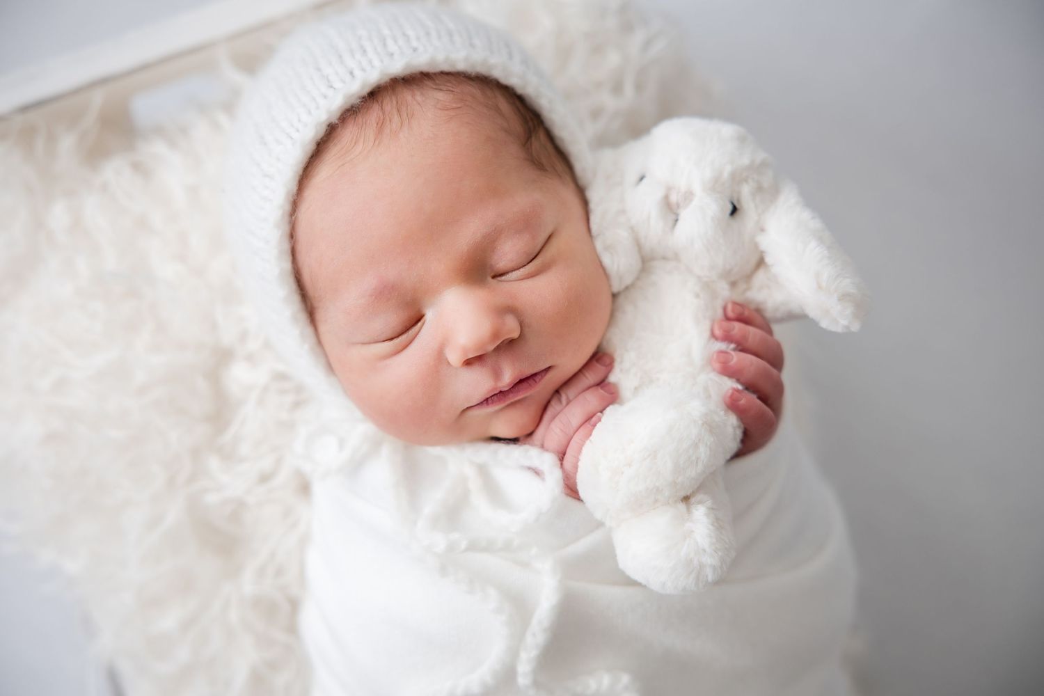 Sleeping newborn baby in white bonnet and swaddle holding a white stuffed bunny toy on a fluffy white blanket.