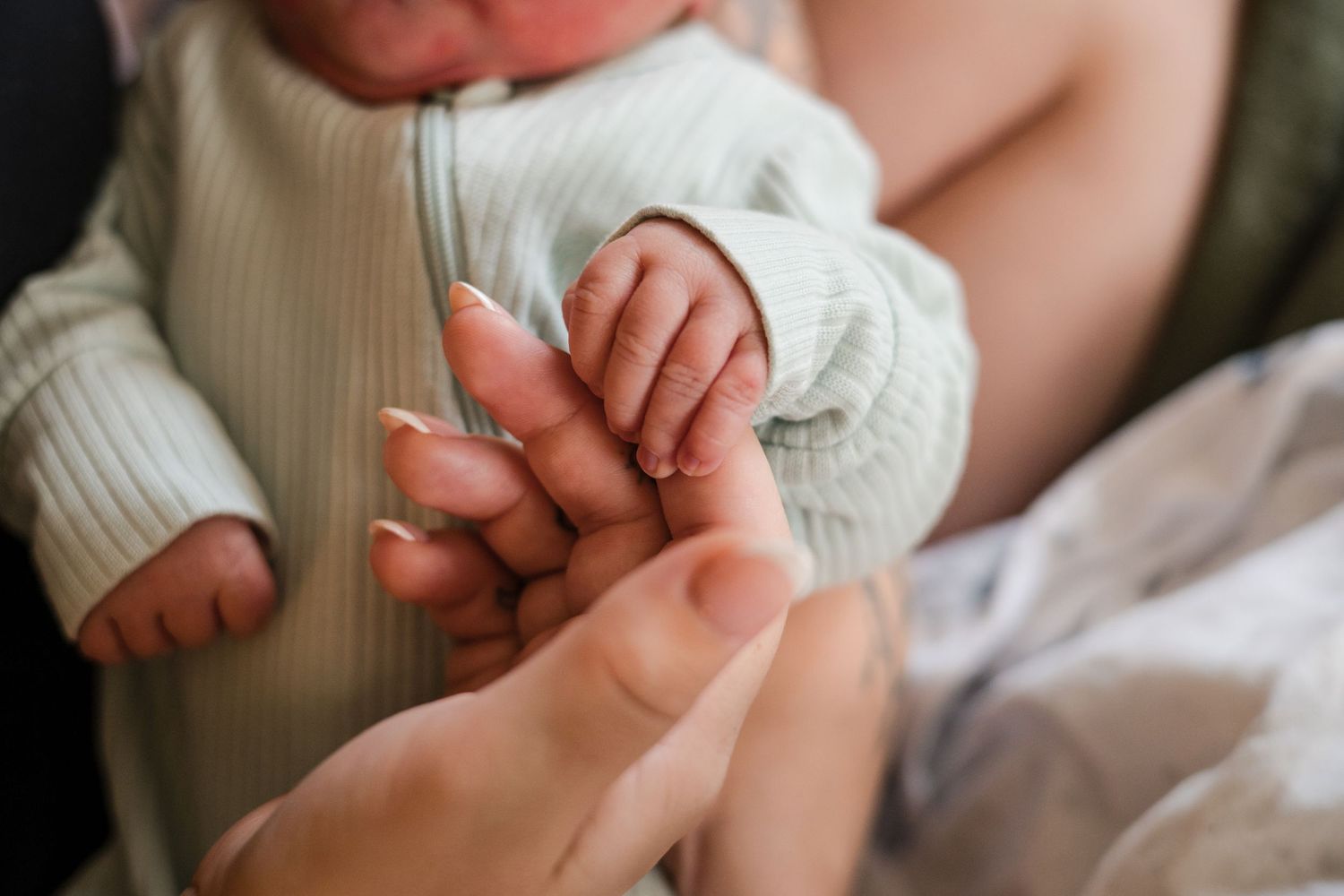 Newborn baby grasping mother's finger, wearing a mint green onesie, in a tender close-up bonding moment.
