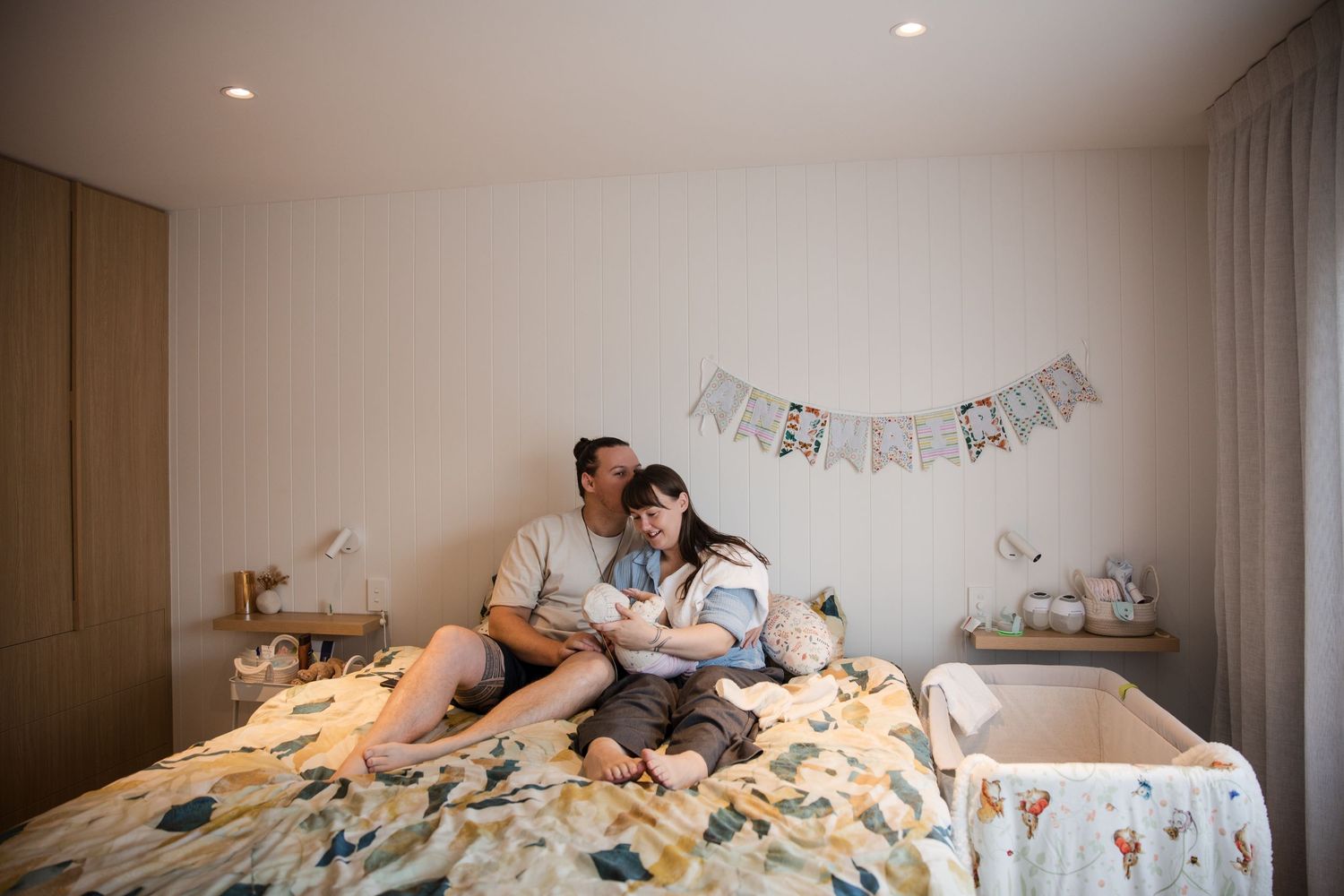 Couple sitting on bed with newborn baby in softly lit nursery room with banner and bedside cot.