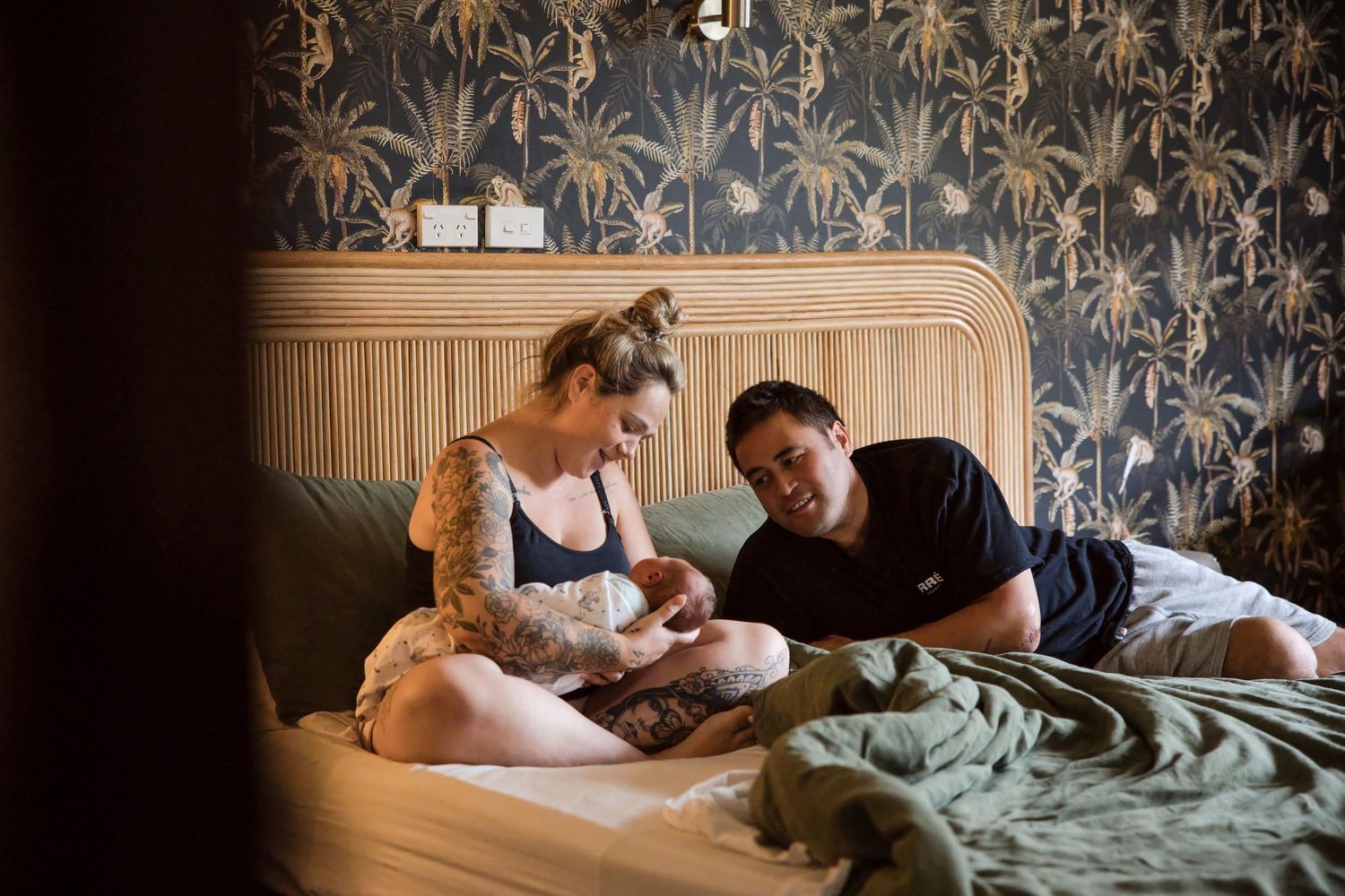 A couple sits on a bed with tropical wallpaper, lovingly gazing at their newborn baby in a cozy bedroom.