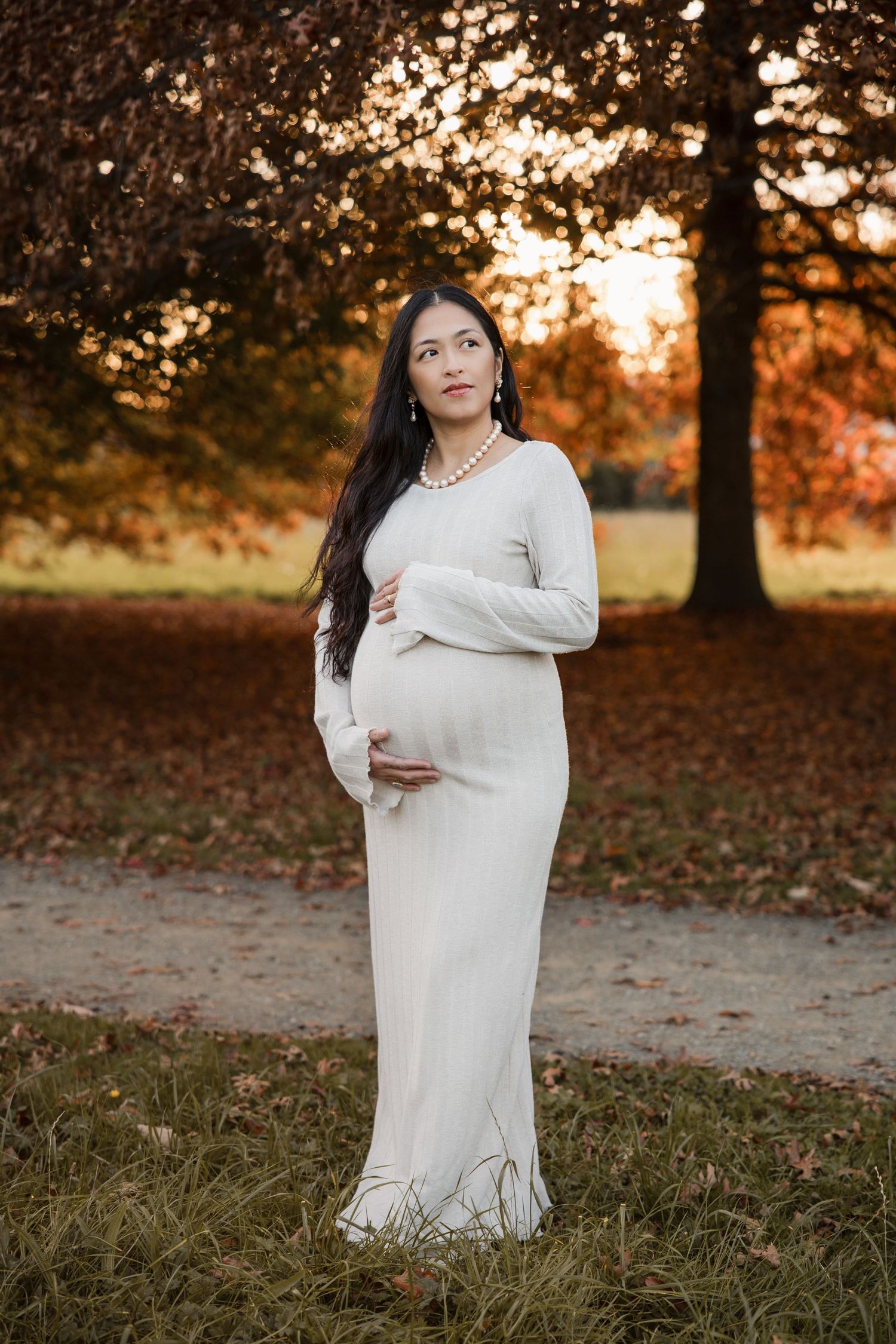 Pregnant woman in white dress cradling belly at golden hour in autumn park with colorful foliage.