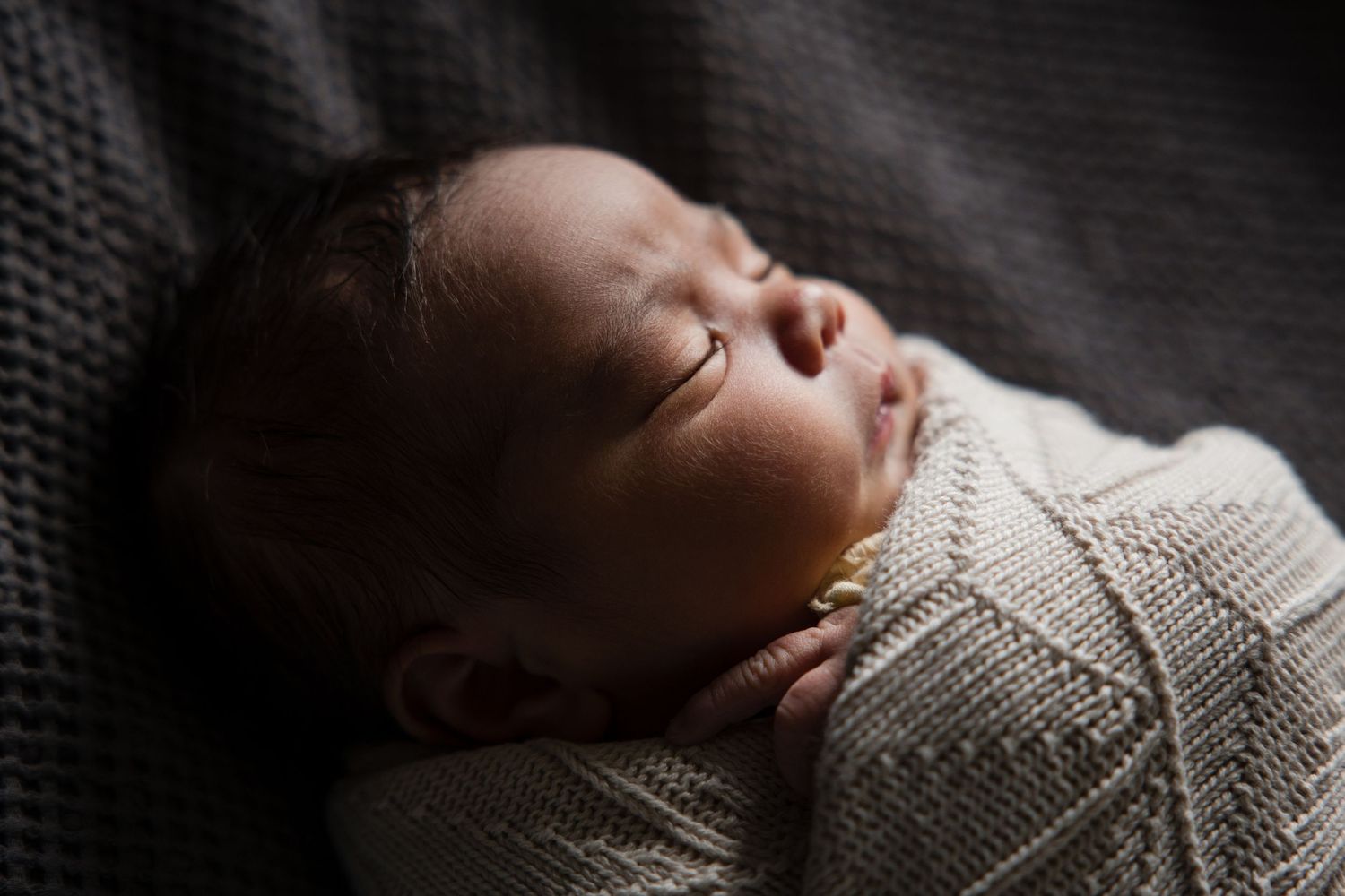 Peaceful newborn baby sleeping wrapped in a soft cream knit blanket, captured in a serene dark moody portrait.