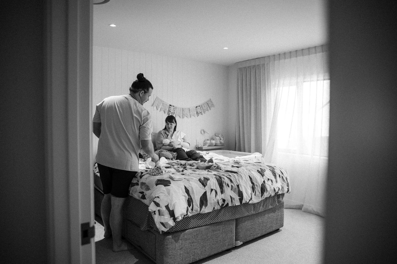 Black and white photo of a mother and child on a bed in a bright bedroom.
