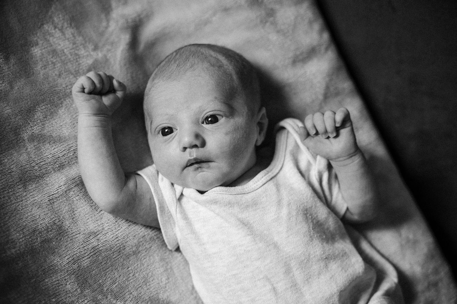 Black and white photo of a newborn baby lying on a blanket with arms raised, wearing a white onesie.