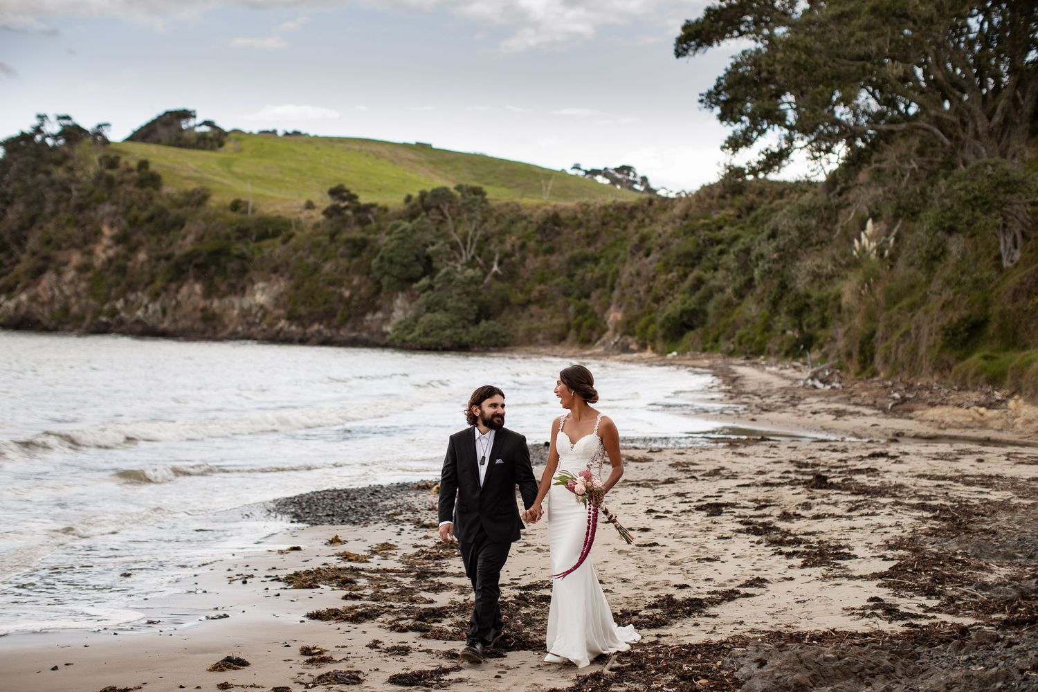 A couple walks along a scenic rocky beach with rolling green hills and cliffs in the background.