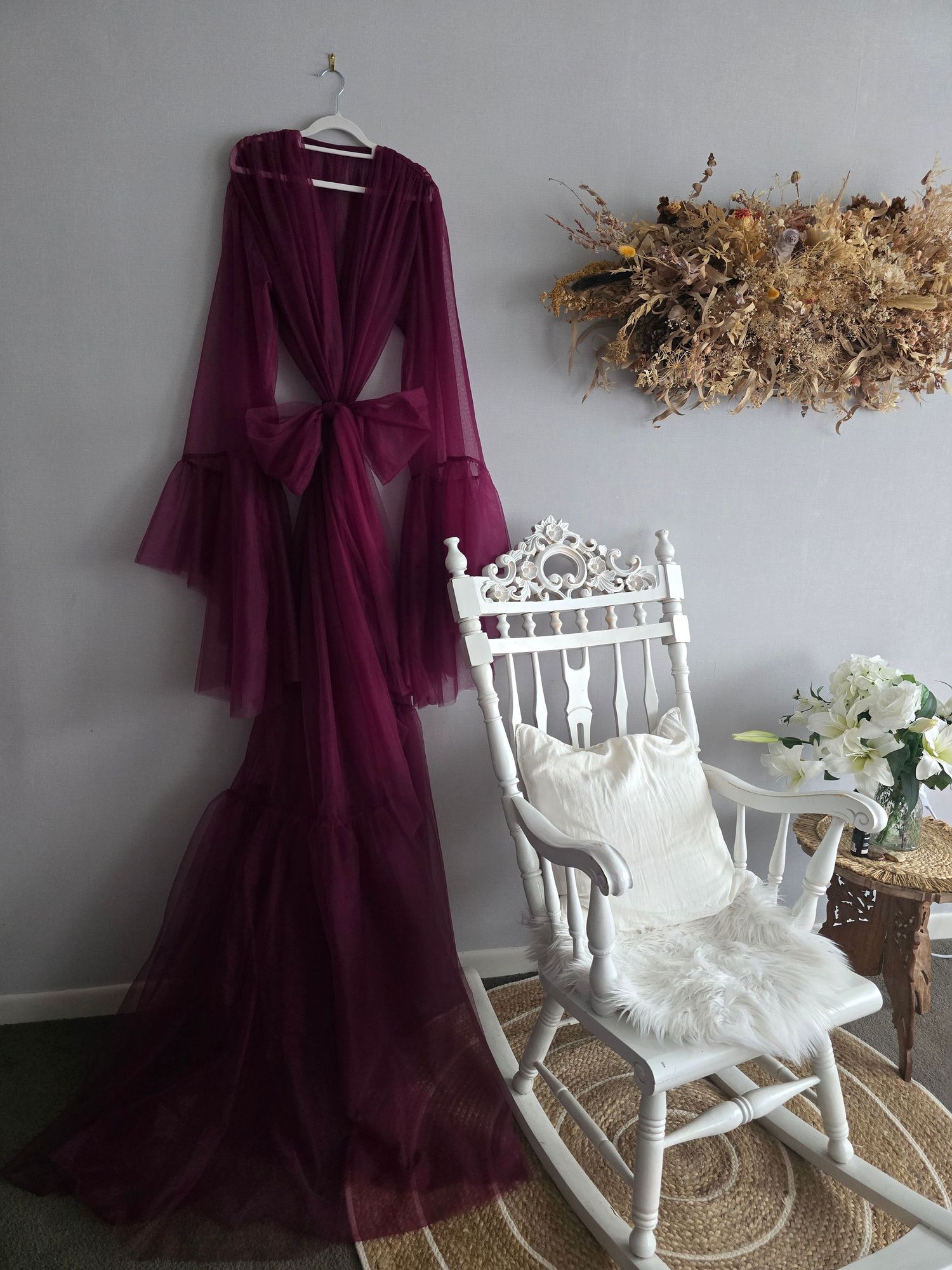 Elegant burgundy tulle robe with bell sleeves displayed on hanger beside white vintage rocking chair with floral decor.