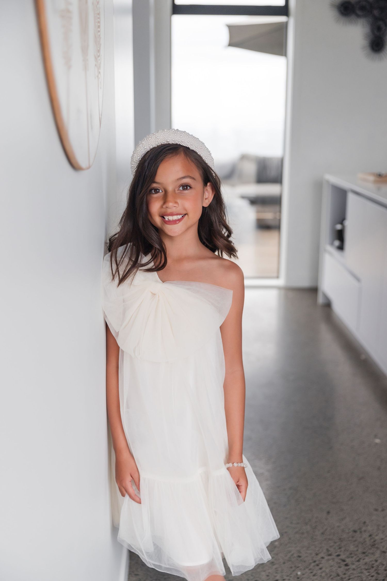 Young person in a flowing white one-shoulder dress stands smiling in a modern interior with polished concrete floors.
