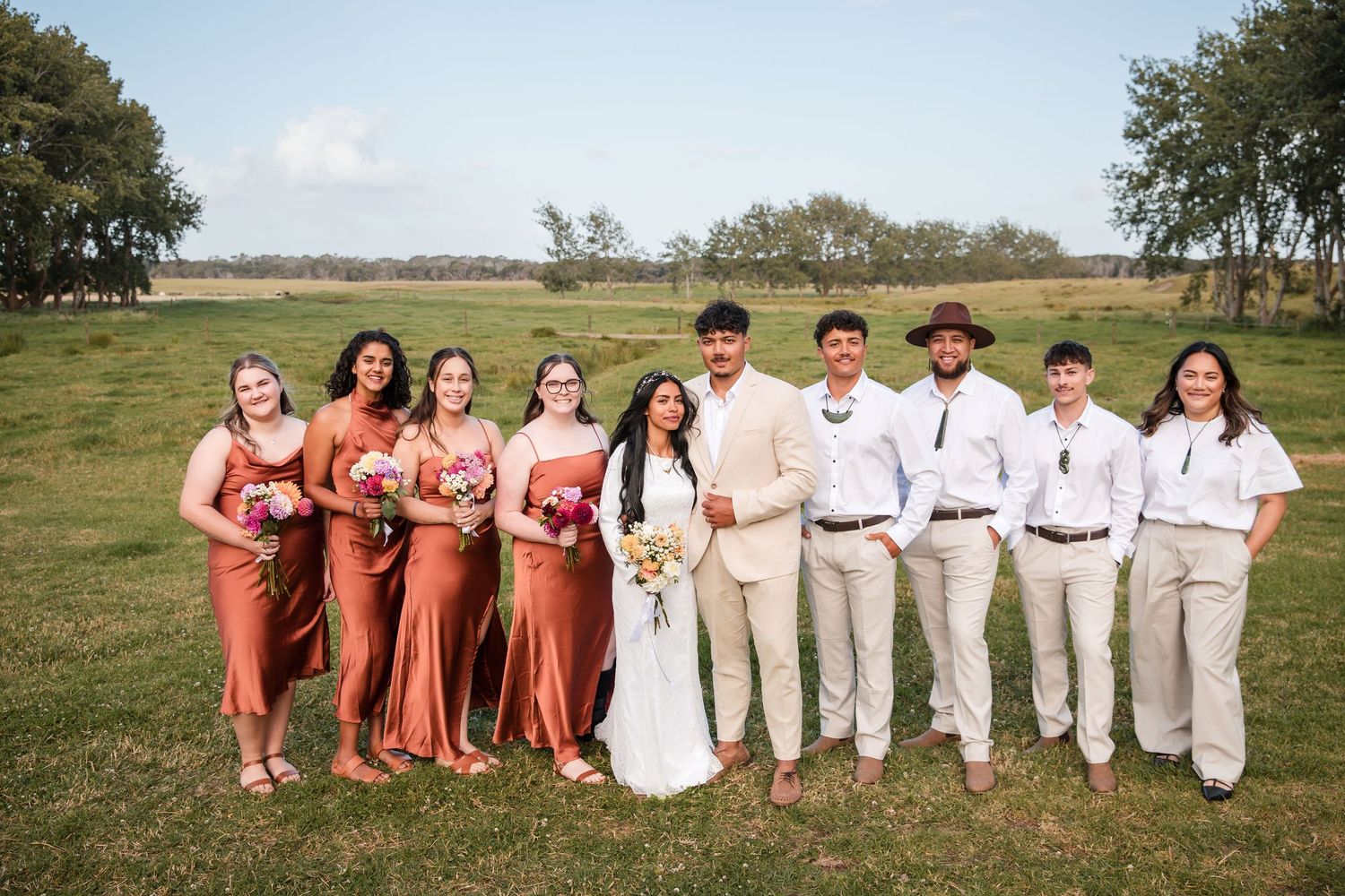Wedding party poses together in a field wearing coral and neutral colored attire on a sunny day.