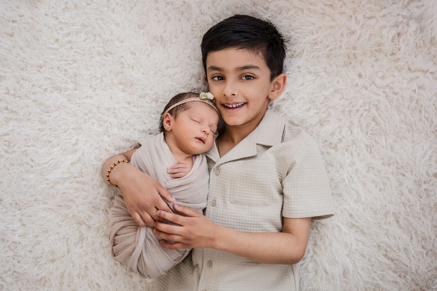 Smiling boy lying on fluffy white rug gently holding a sleeping swaddled newborn baby with a floral headband.
