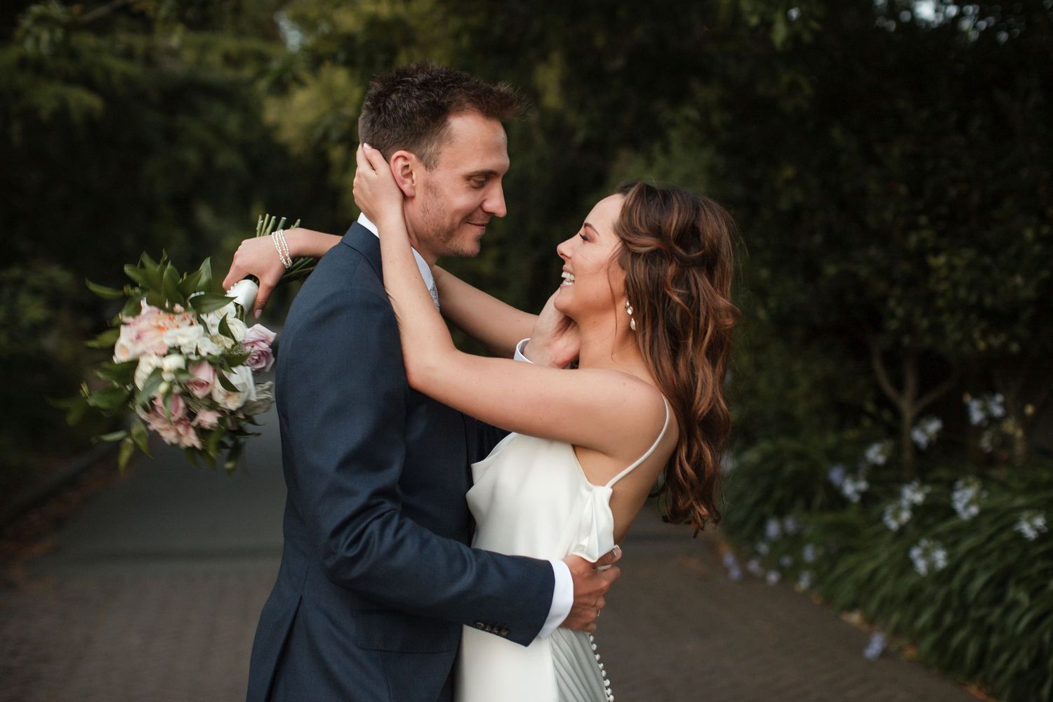 A couple in wedding attire share an intimate embrace while dancing outdoors in a garden setting.