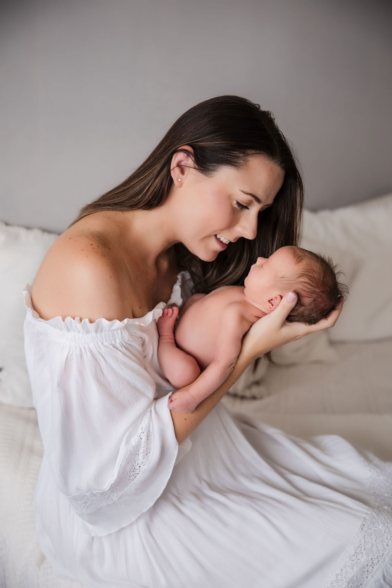 A mother in a white off-shoulder dress tenderly holds her sleeping newborn baby while sitting on a bed.