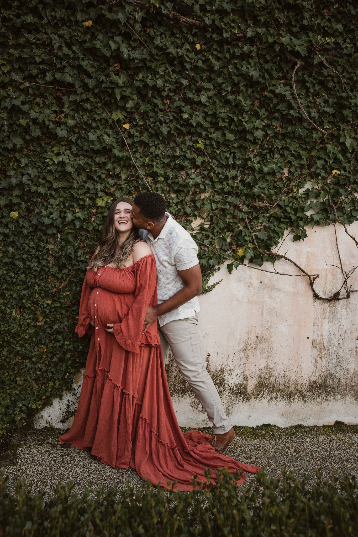 Pregnant woman in flowing rust dress poses with partner against ivy-covered wall for maternity photo shoot.