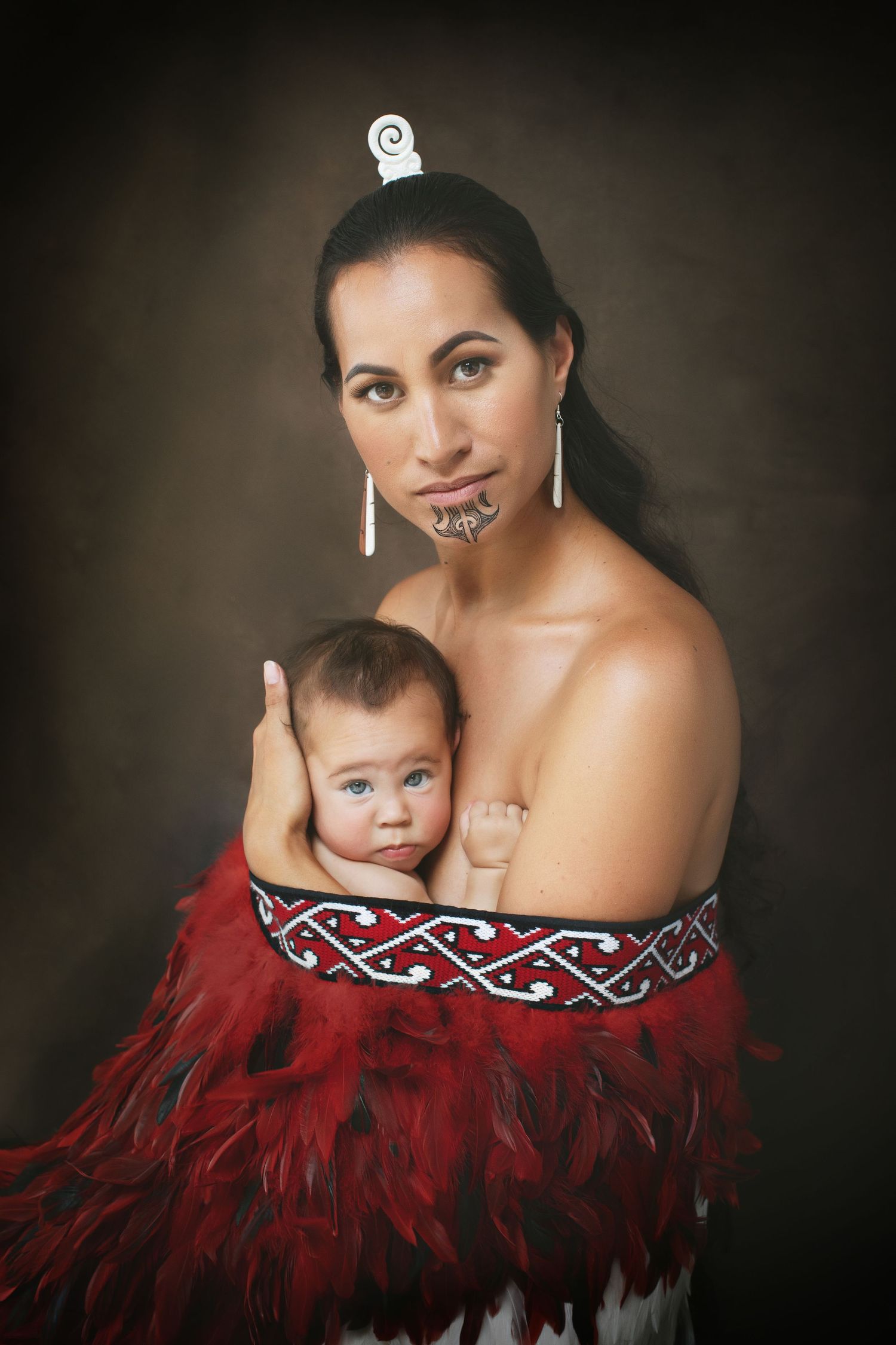 Maori mother with traditional chin tattoo holds baby while wearing red feathered cloak. Cambridge Waikato NZ Photographer