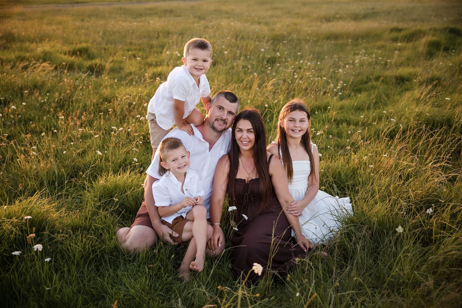 Family of five posing in a sunny green meadow with wildflowers, parents seated with three children smiling warmly.