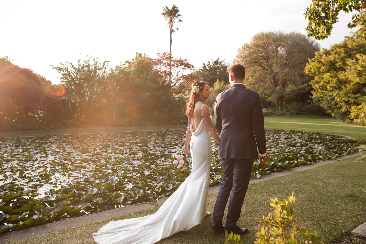 A couple in wedding attire stands at sunset by a pond with lily pads and a palm tree in the background.