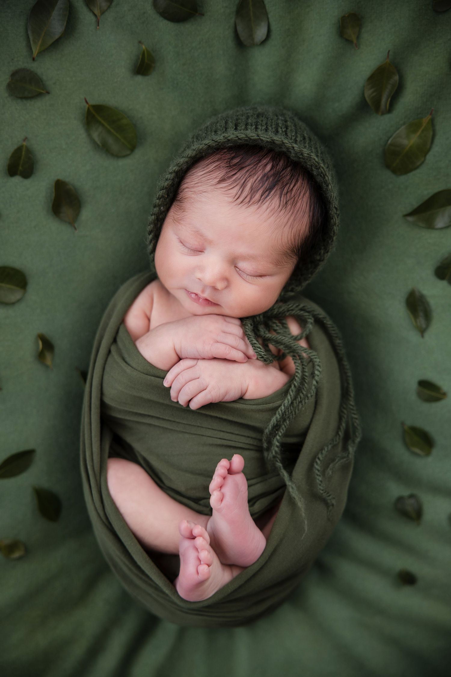 Sleeping newborn baby wrapped in olive green fabric against a dark green textured background.