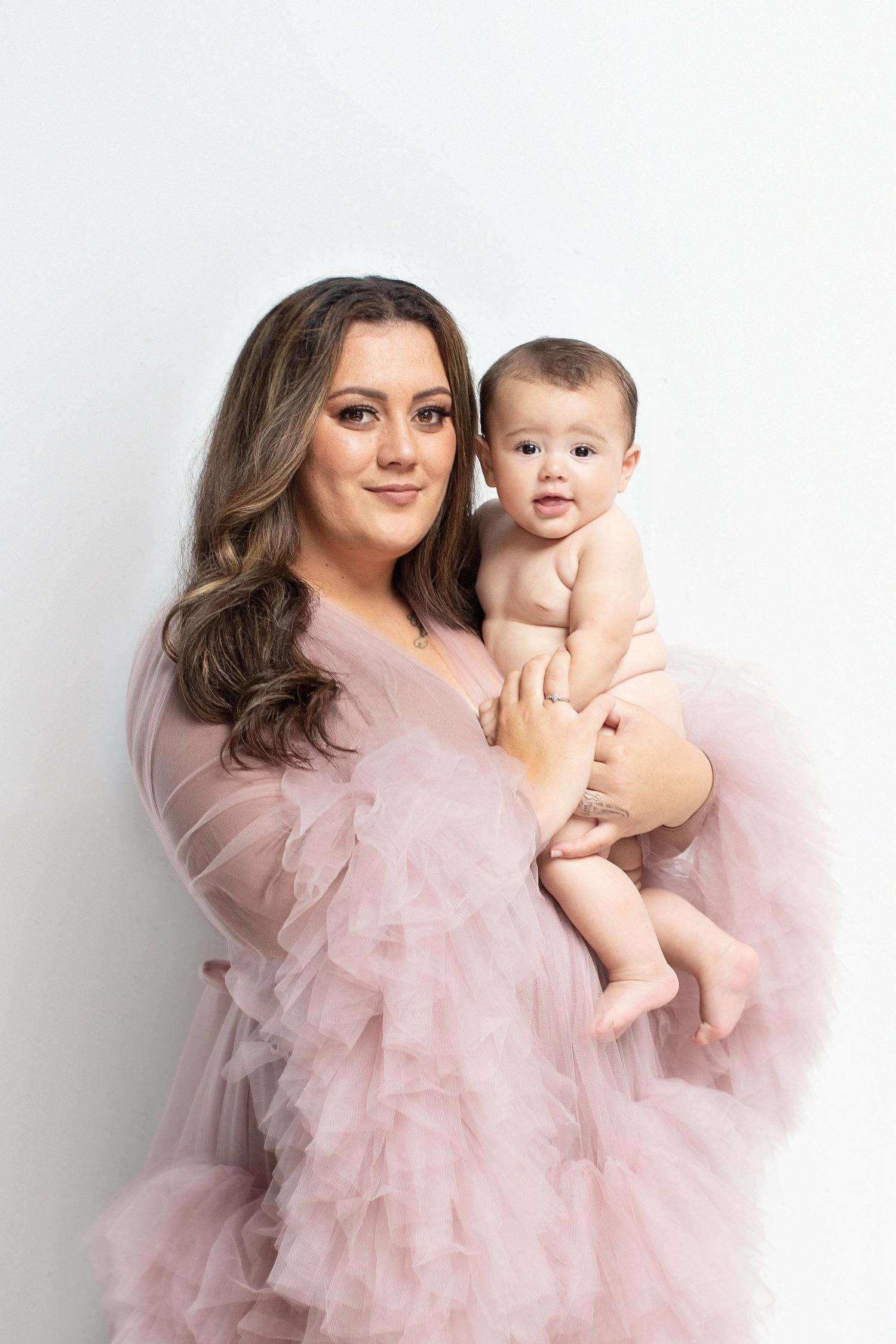 A woman in a pink tulle robe holds her baby infant during a professional studio portrait session.