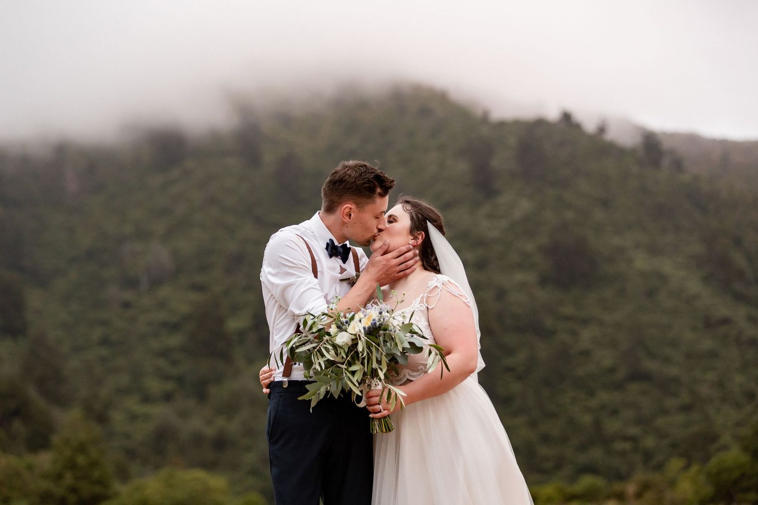 A romantic wedding couple shares an intimate moment in front of misty mountain scenery while holding a green bouquet.
