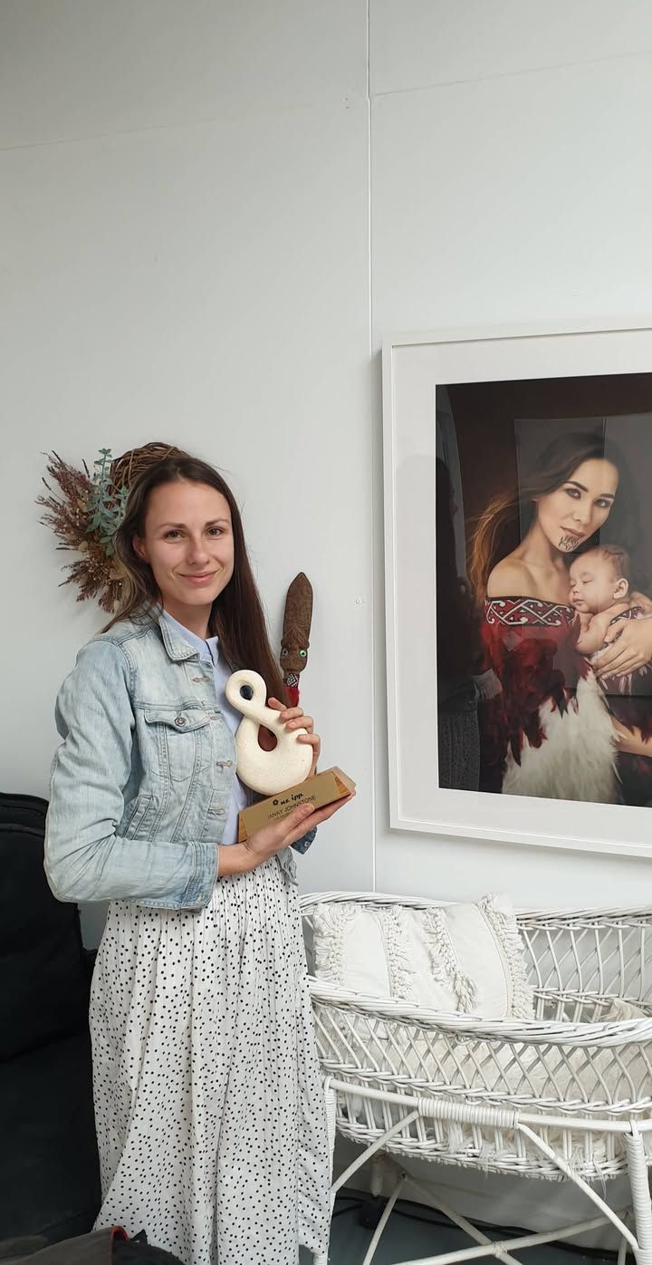 Person in denim jacket holding a plush toy while standing near a white bassinet in a gallery-like setting.