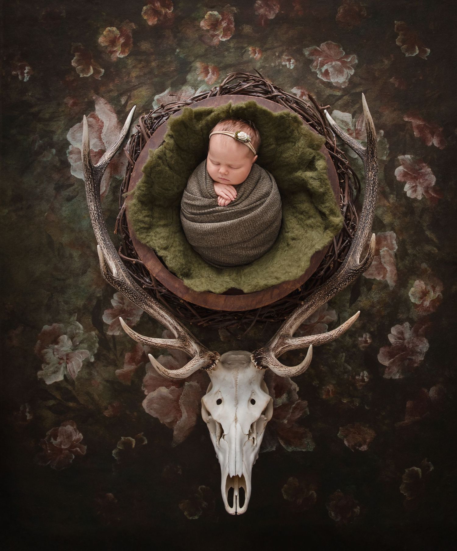 Newborn baby wrapped in gray swaddle resting in a nest atop a deer skull, surrounded by floral backdrop.