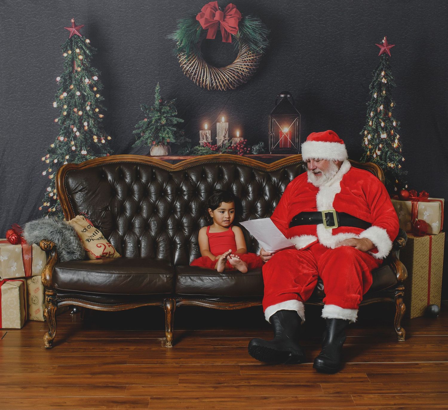 Santa Claus sits on a vintage leather couch next to a child with decorated Christmas trees and wreaths in background.