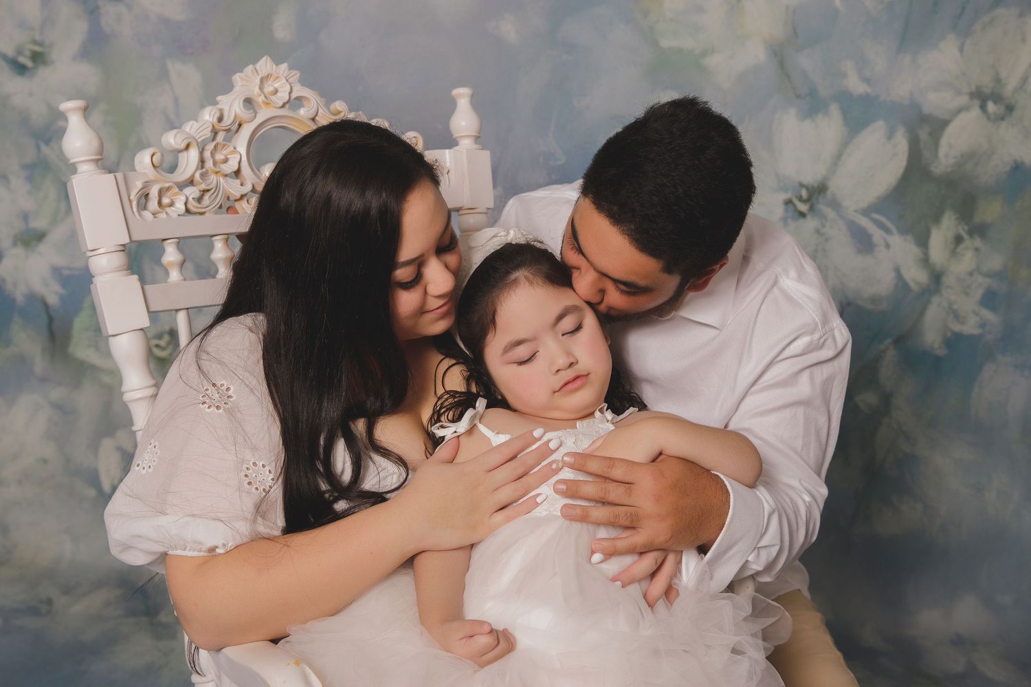 A tender family moment as parents embrace their child while sitting in an ornate white chair against a muted floral backdrop.