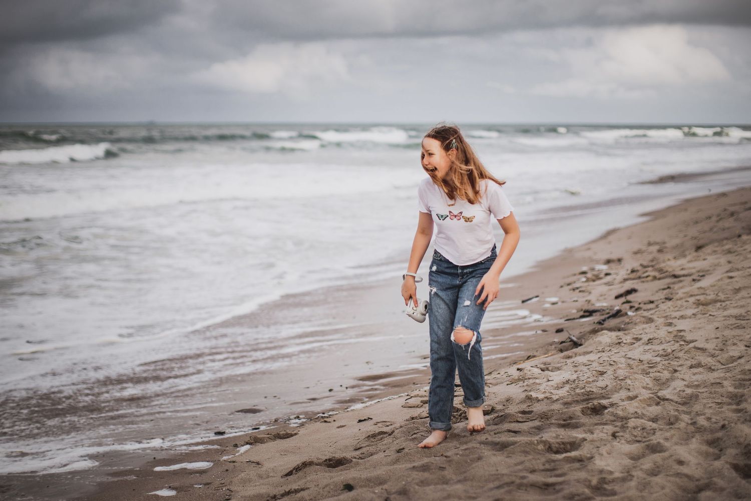 Teenage girl walking barefoot along stormy beach wearing casual denim and white top.