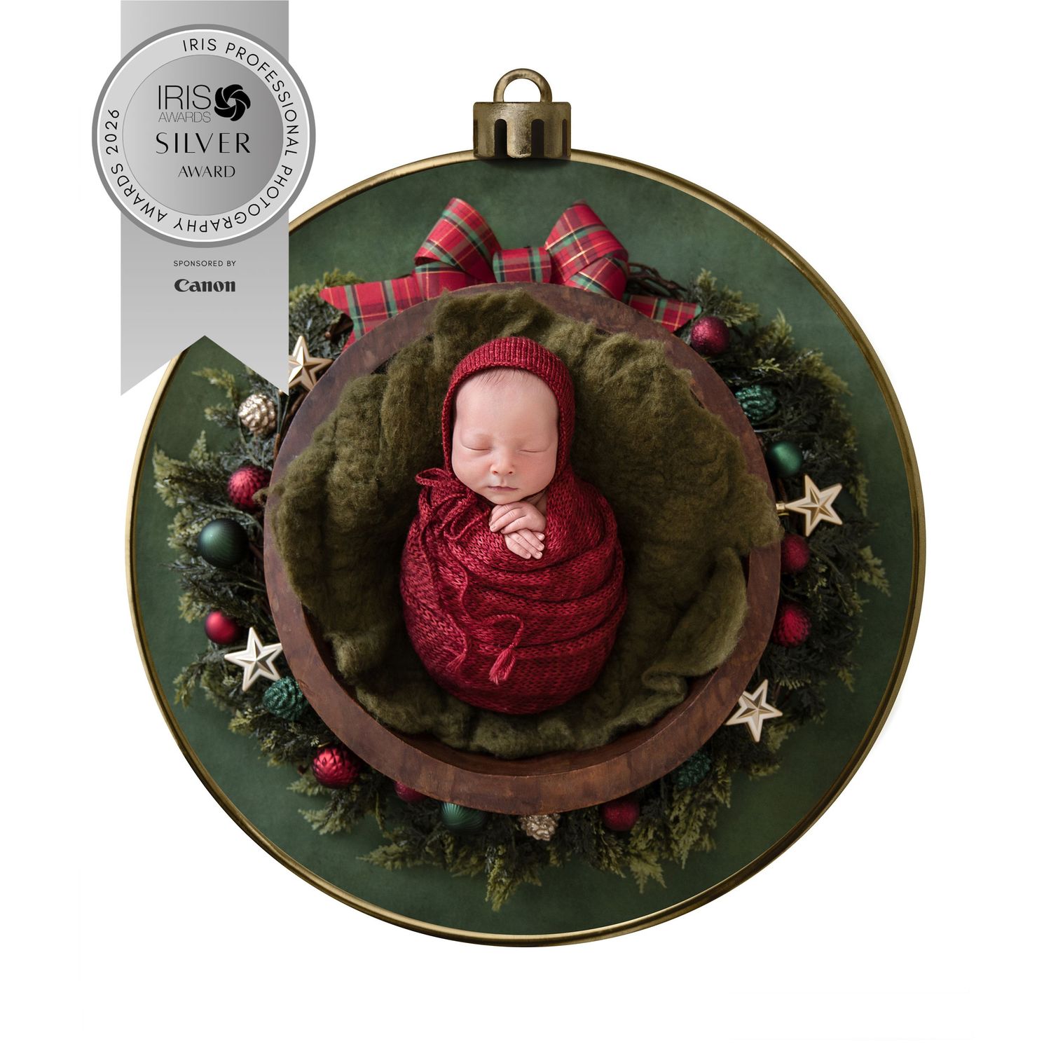 Award-winning newborn photo of sleeping baby in red wrap inside Christmas ornament with festive greenery and bow decoration.