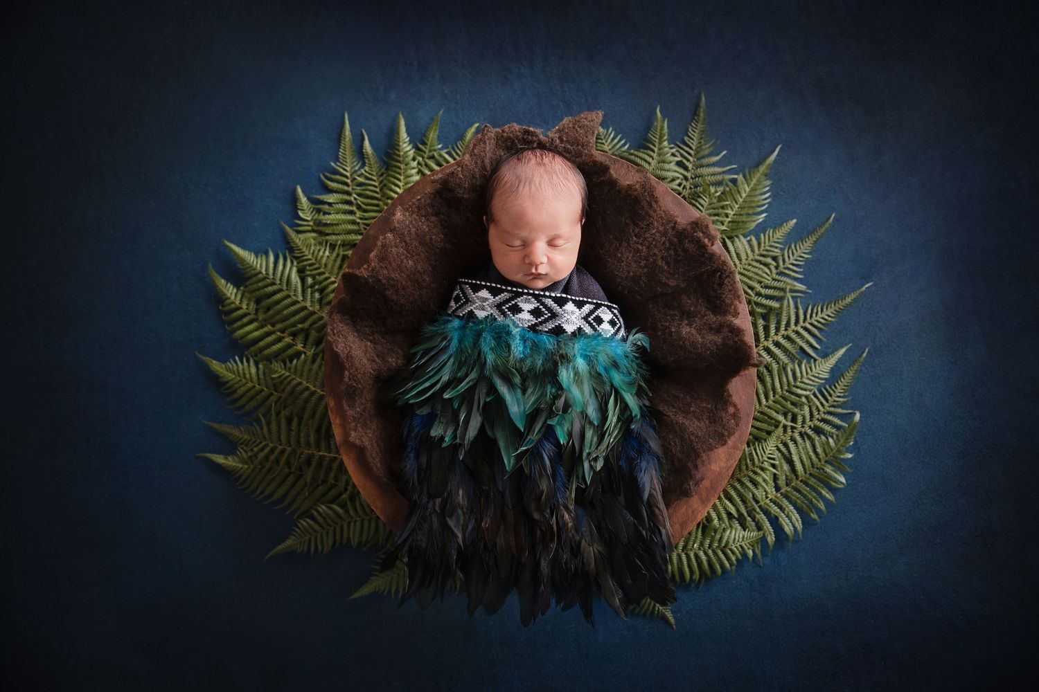 Newborn baby wrapped in teal feathers and tribal blanket, nestled in brown fur bowl with ferns on blue background.