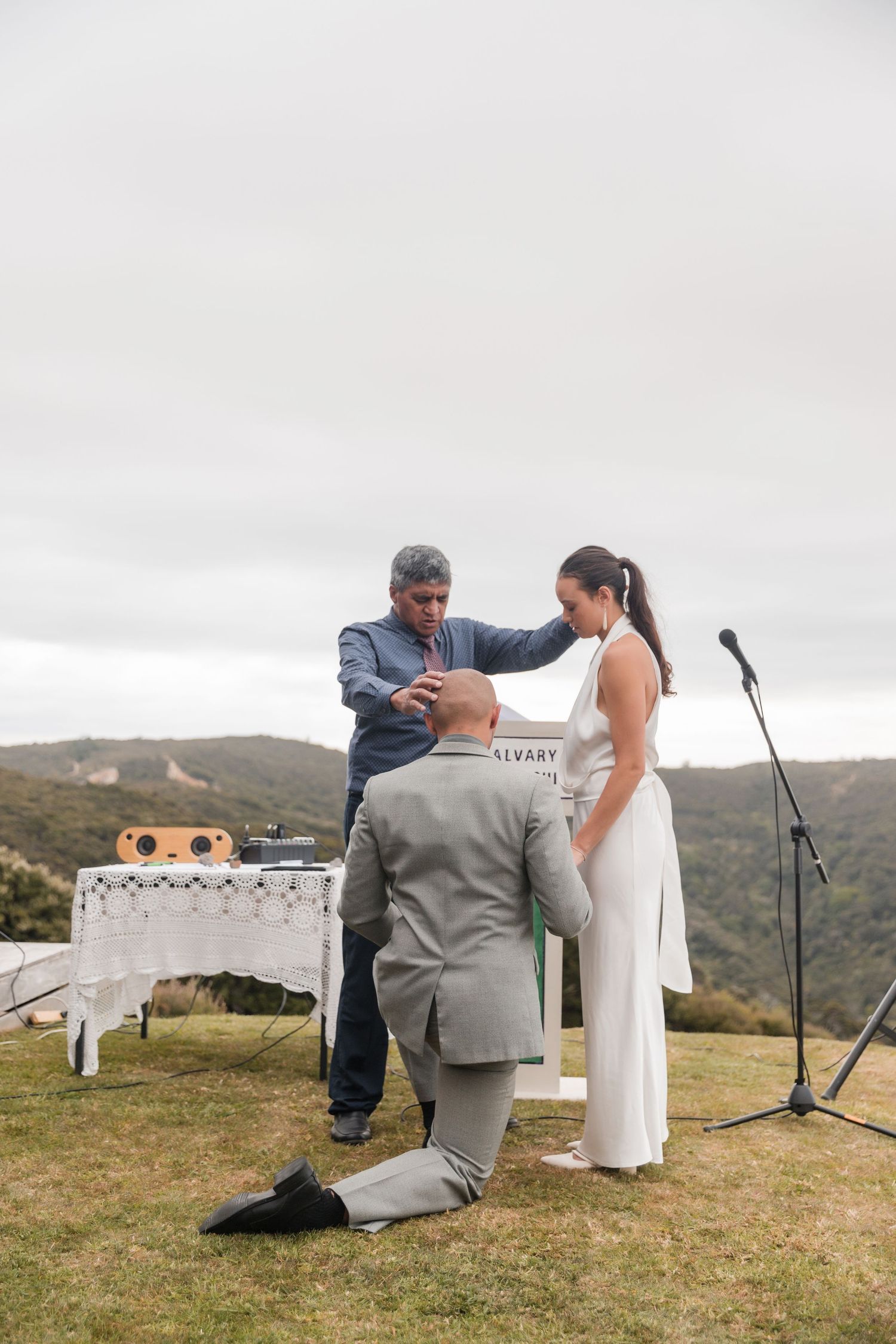 Wedding ceremony taking place outdoors on a hillside with an officiant and couple during a cloudy day.