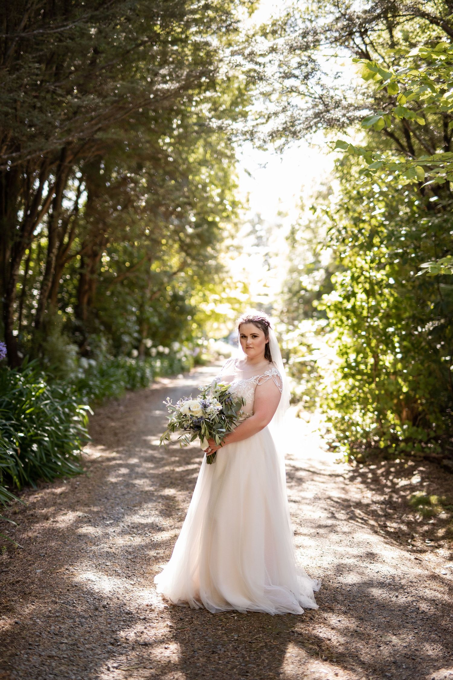 A bride in a flowing white gown poses gracefully on a sunlit forest path surrounded by lush greenery.