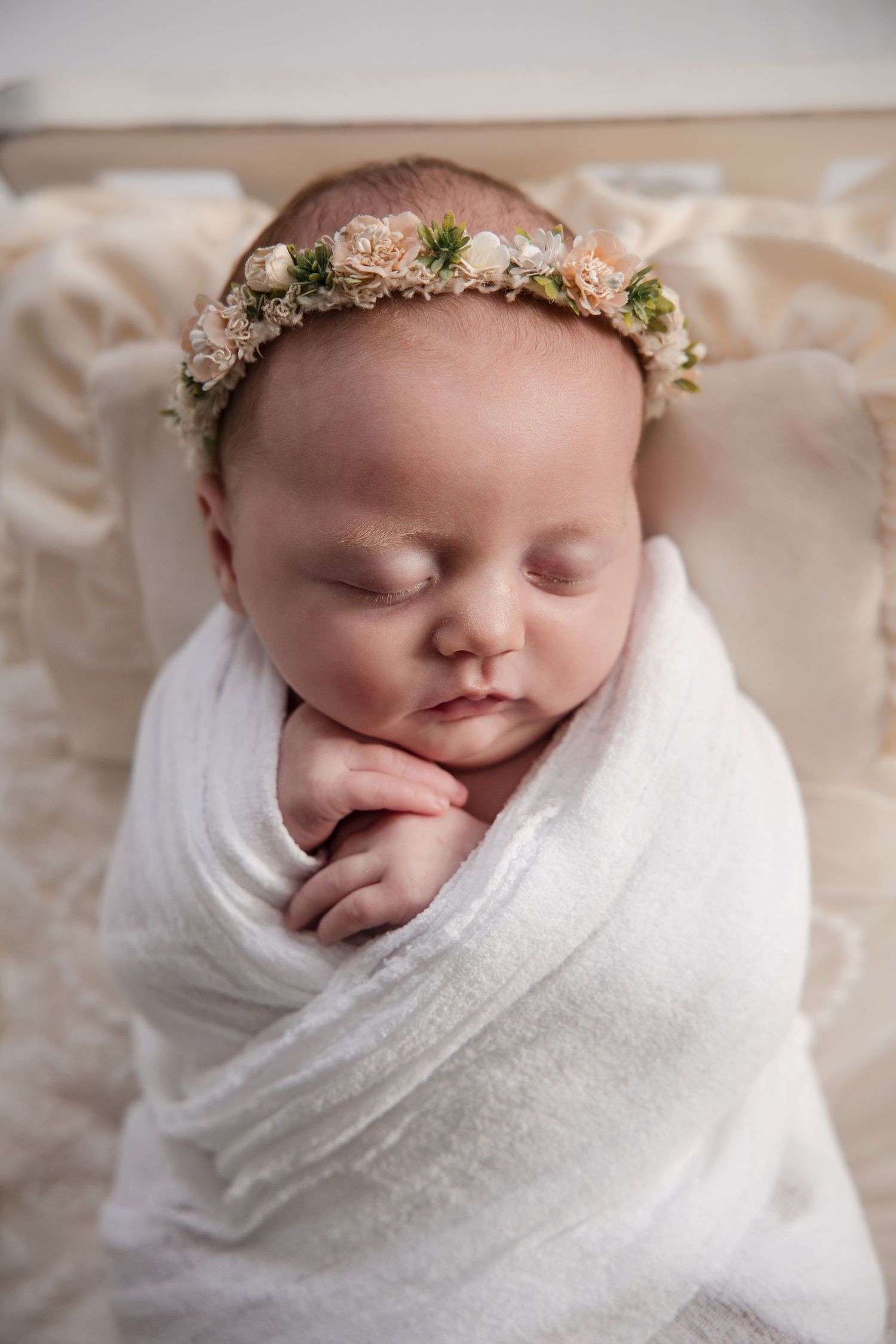Sleeping newborn baby swaddled in white wrap wearing a delicate floral crown, peacefully resting on a soft blanket.