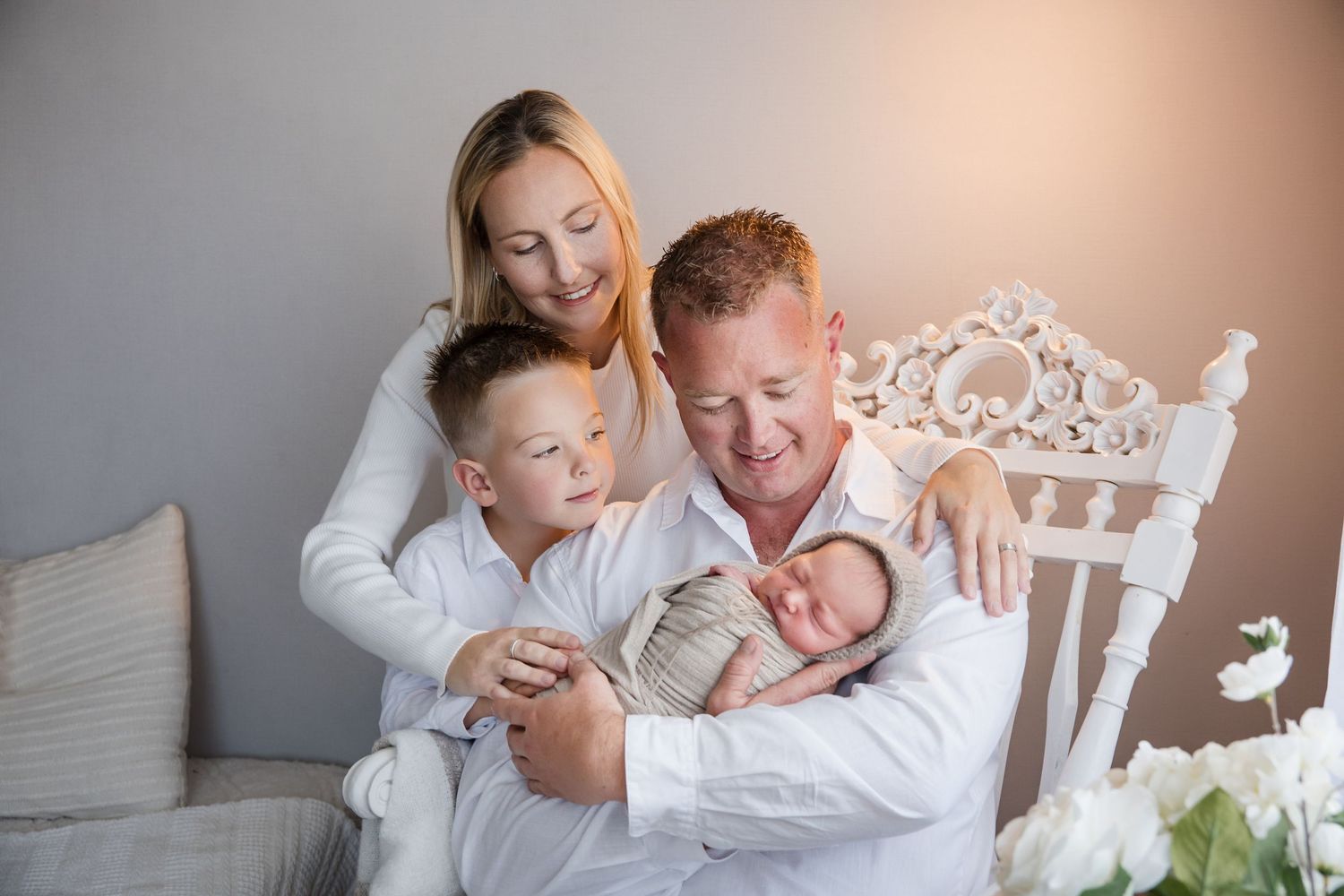 Happy family of four posing for newborn photo, parents with young son holding sleeping baby in white studio setting.