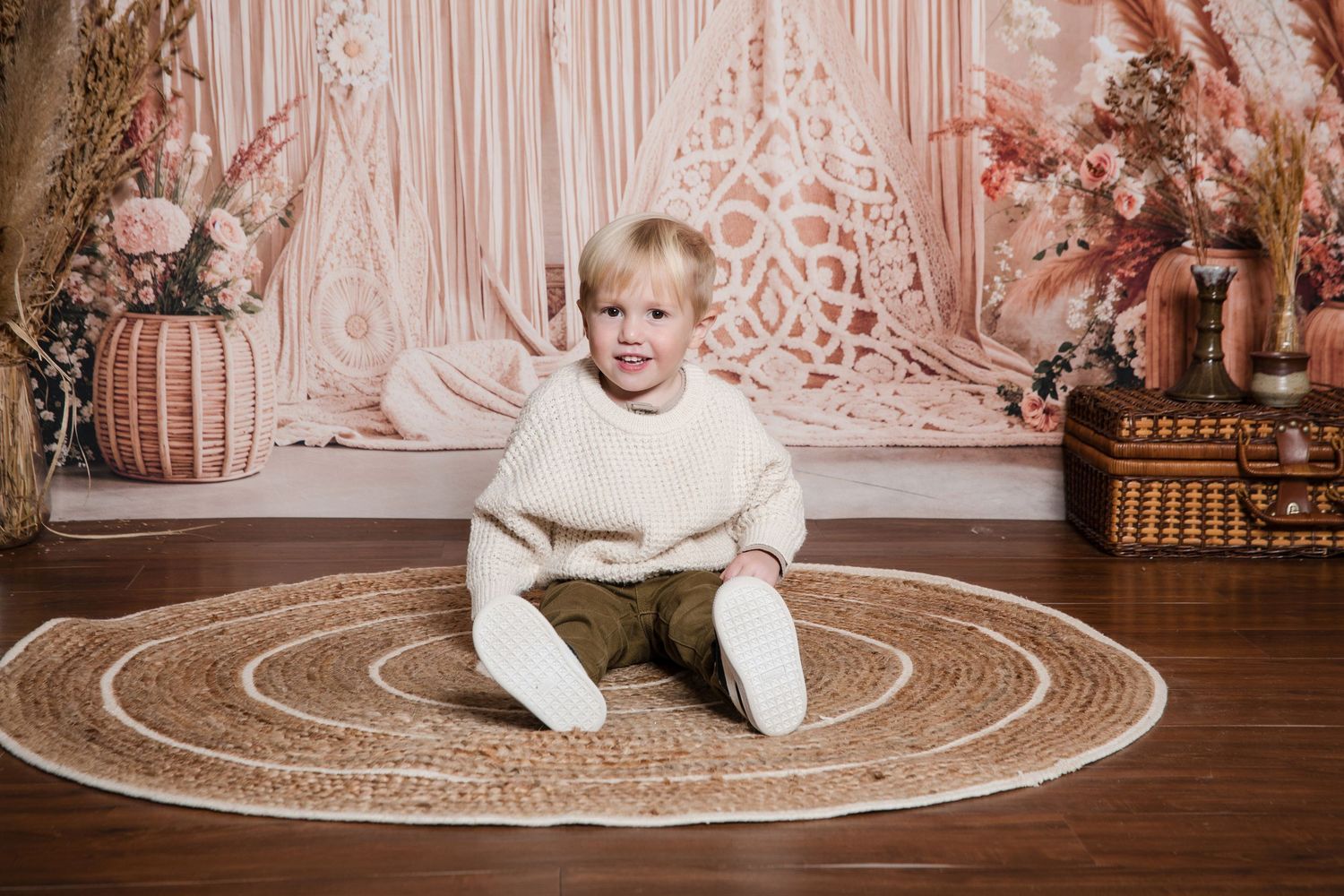 A baby in cream-colored clothing sits on a round rug against a pink floral backdrop with vintage decor.