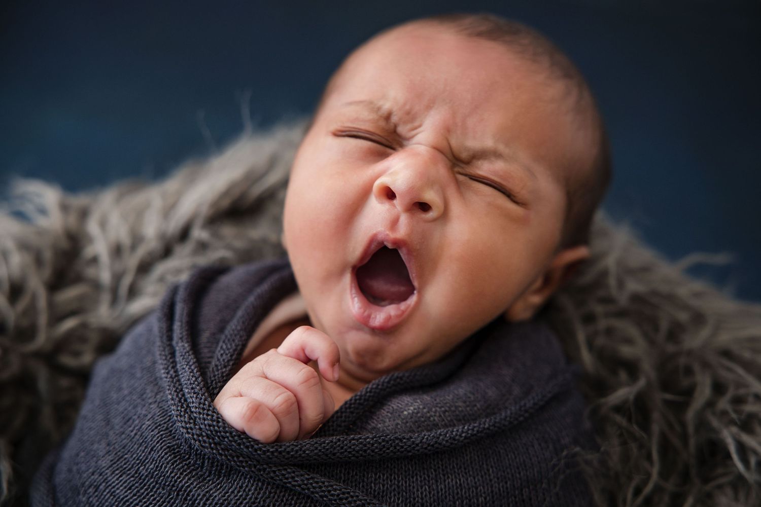 Sleeping newborn baby in gray wrap yawns while lying on textured gray blanket against dark backdrop.