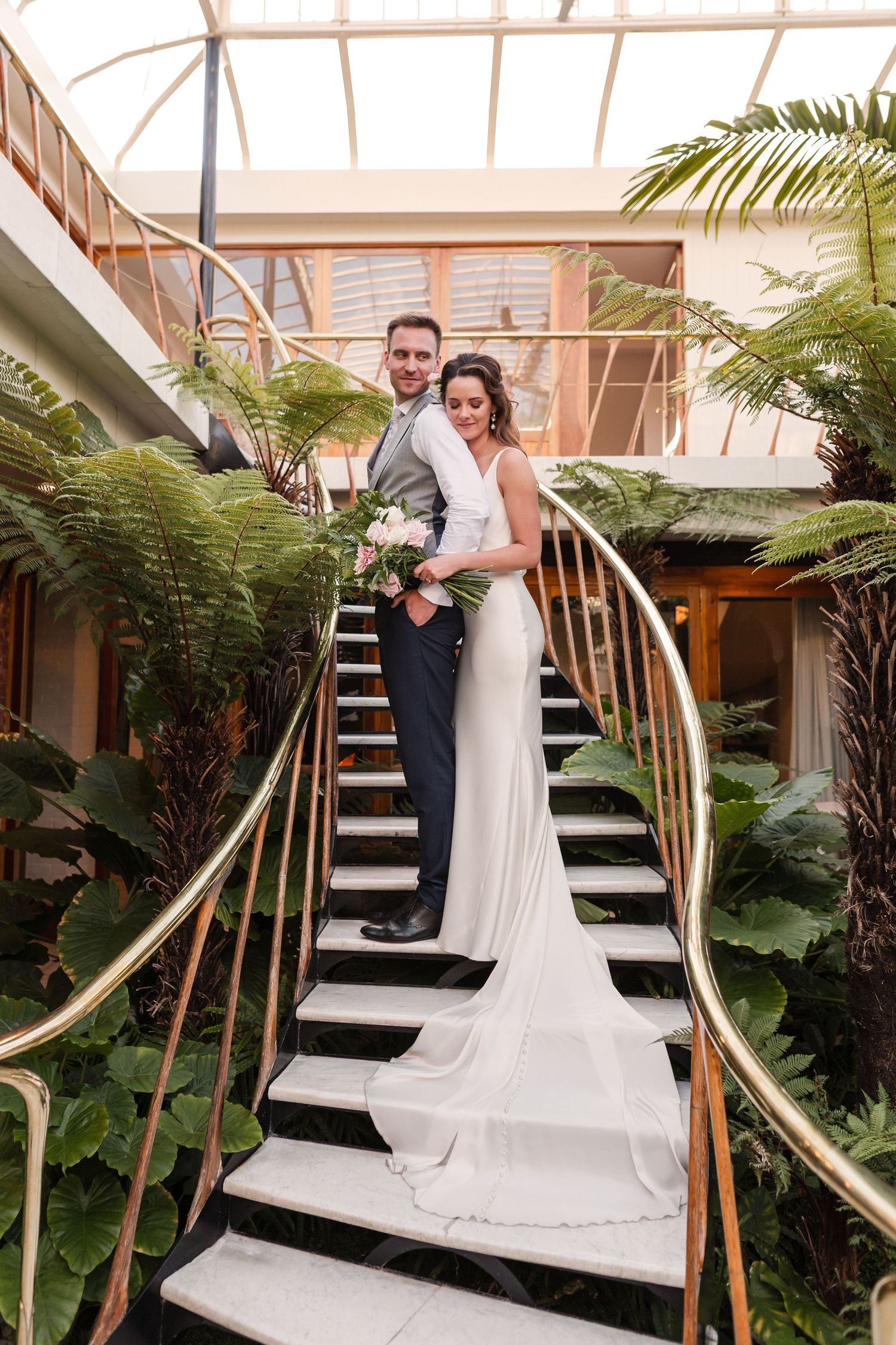 A couple shares an intimate moment in a modern indoor venue with curved bamboo arches and tropical ferns.