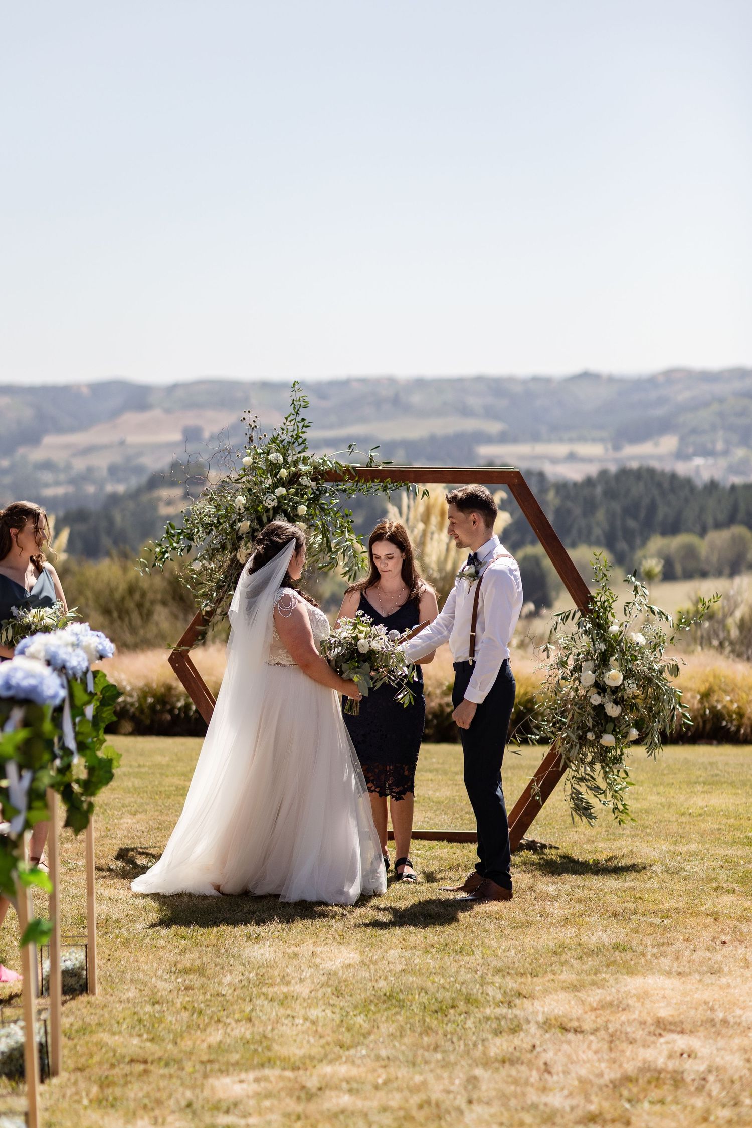 Wedding ceremony guests gather around a hexagonal wood arch decorated with floral arrangements on a scenic hillside.