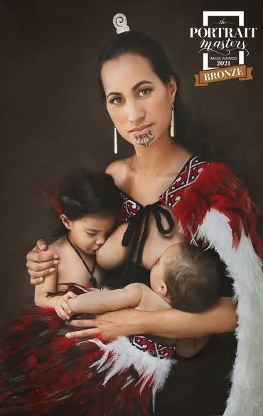 Mother in traditional Maori attire nursing two children while wearing cultural face markings and feathered cloak.