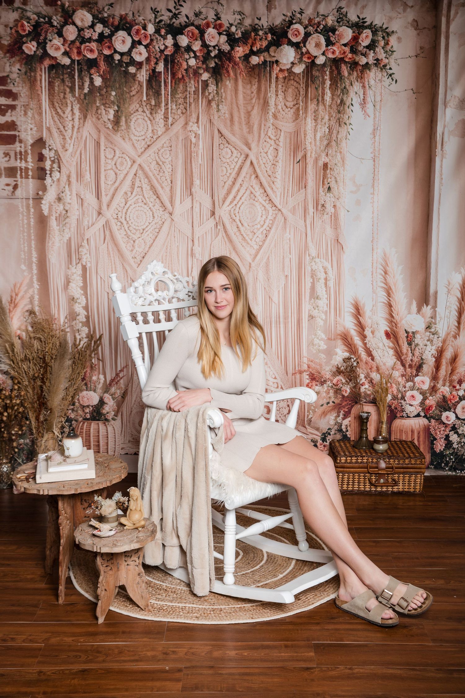 Boho-style photo setting with macrame backdrop, floral arch, rocking chair, and pink and neutral decor elements.