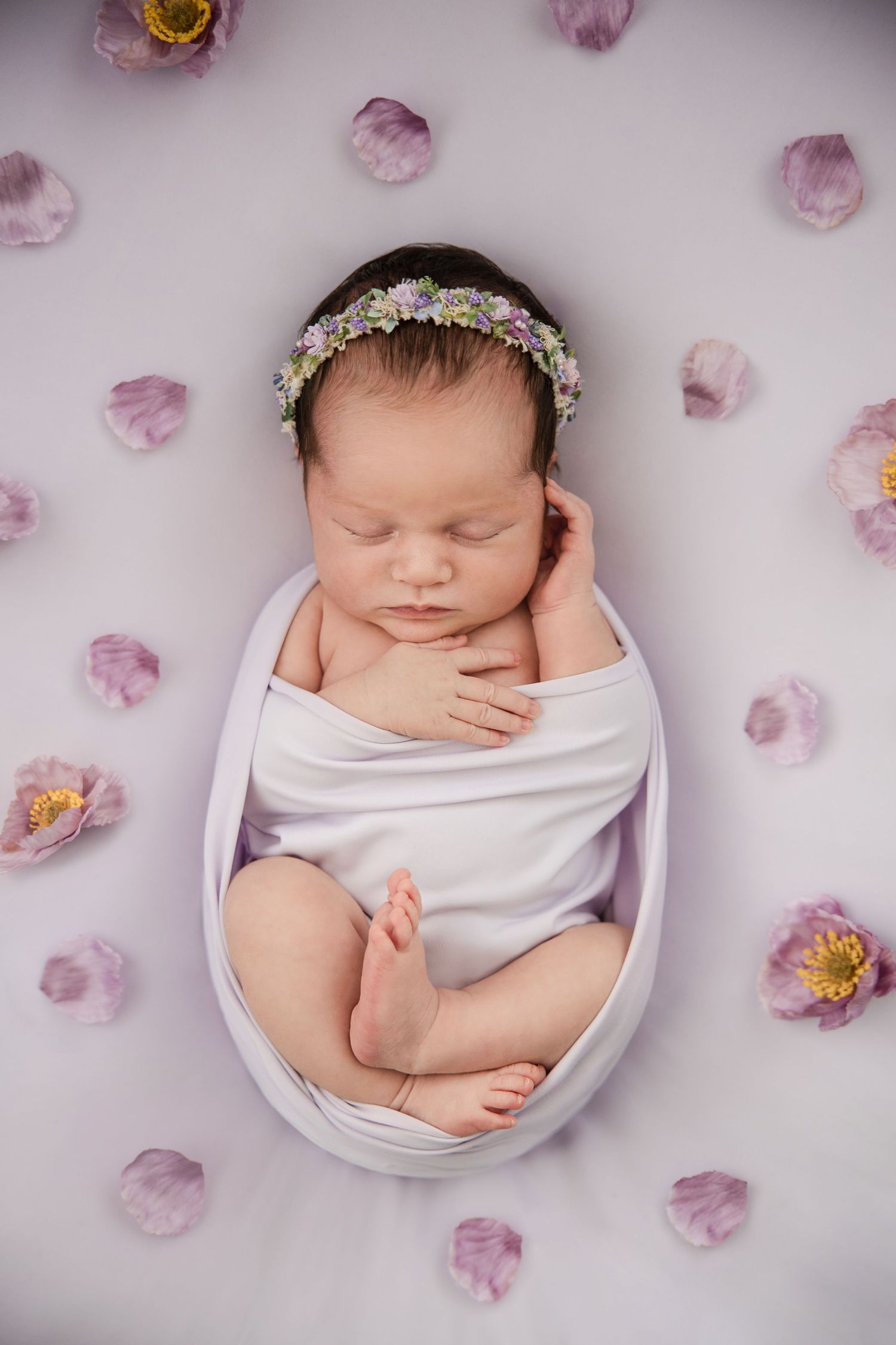 Sleeping newborn wrapped in white fabric surrounded by delicate pink flower petals in artistic portrait series.
