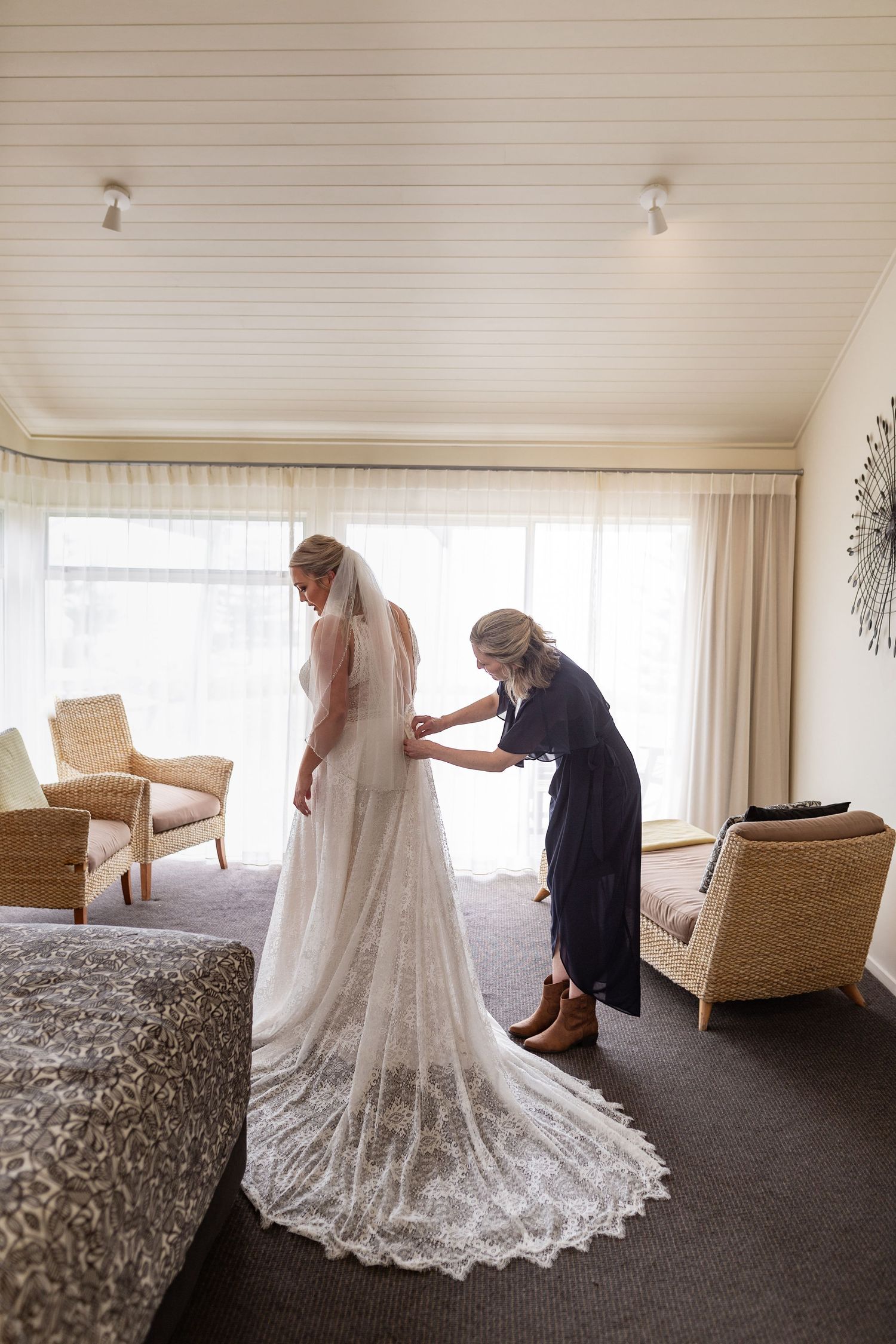 Final adjustments being made to a beautiful lace wedding gown with a long flowing train in a sunlit room.