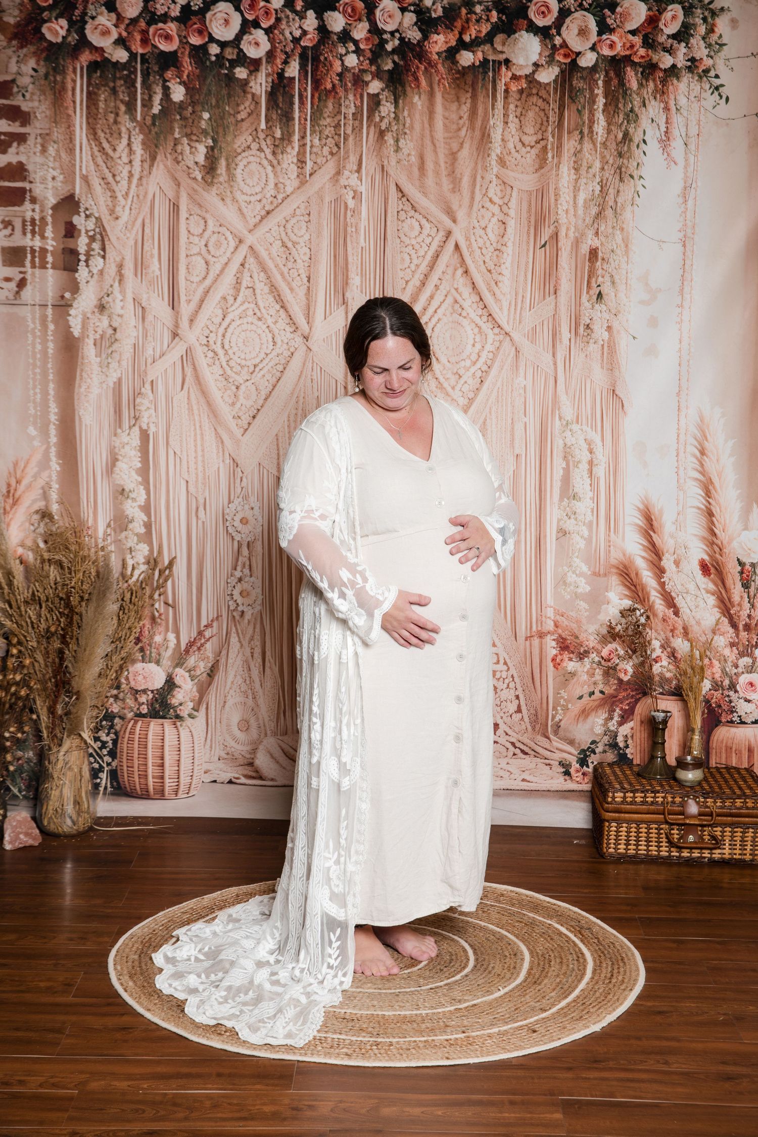 A maternity photo features a woman in a flowing white lace dress against a macrame backdrop with floral decor and pampas grass.