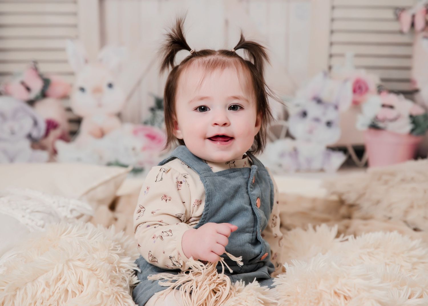 Baby in denim overalls sits on fluffy rug with pink floral backdrop and pigtail hairstyle.
